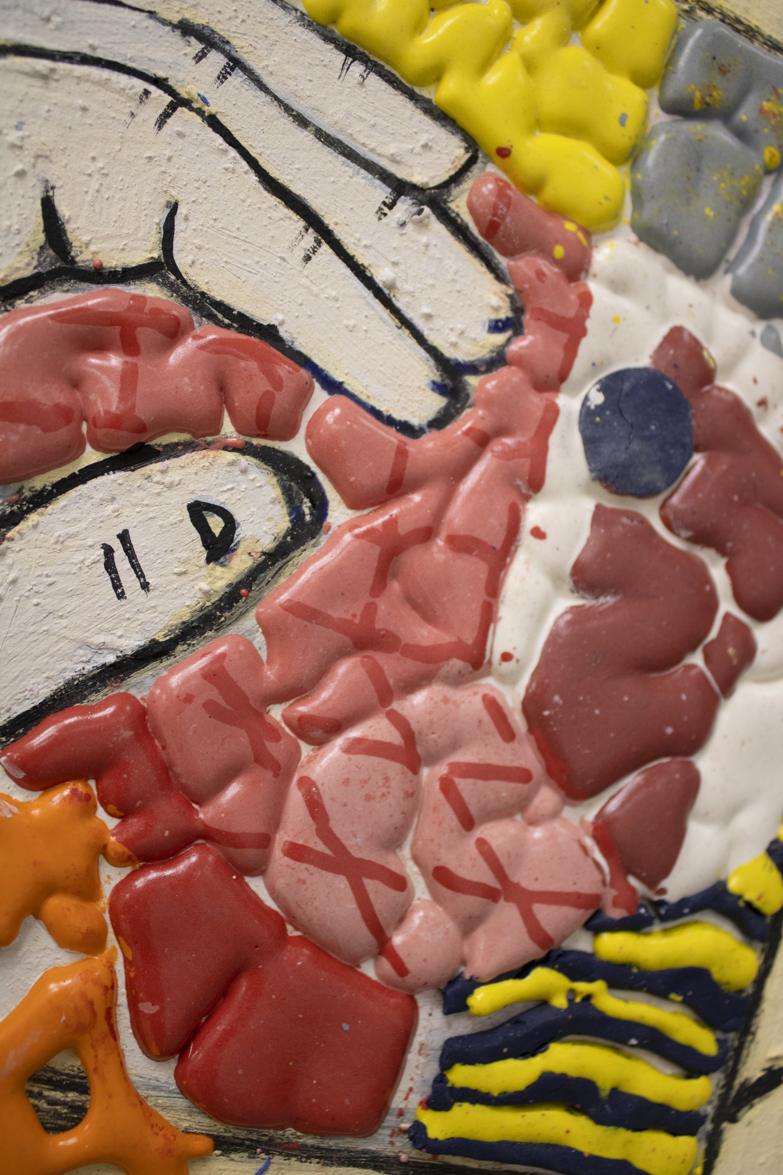 Close-up of a colorful, textured artwork featuring painted and shaped clay or ceramic pieces in various colors including orange, pink, yellow, gray, and dark blue, with visible brush strokes and raised surfaces.