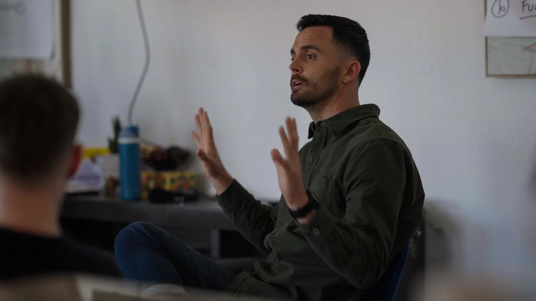 A man with dark hair and wearing a dark shirt is talking and gesturing with his hands while seated in a classroom or meeting room. There is a person with short hair in the foreground, and posters and objects are visible in the background.