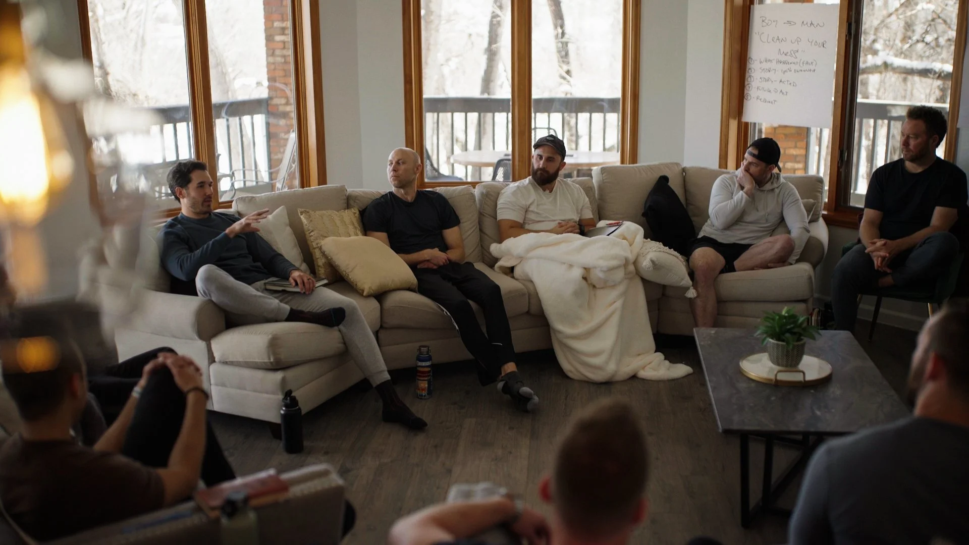 A group of five men sitting on a beige sectional sofa in a living room during a discussion or meeting. There are large windows behind them showing an outdoor balcony with trees. A whiteboard with notes is visible on the right side.