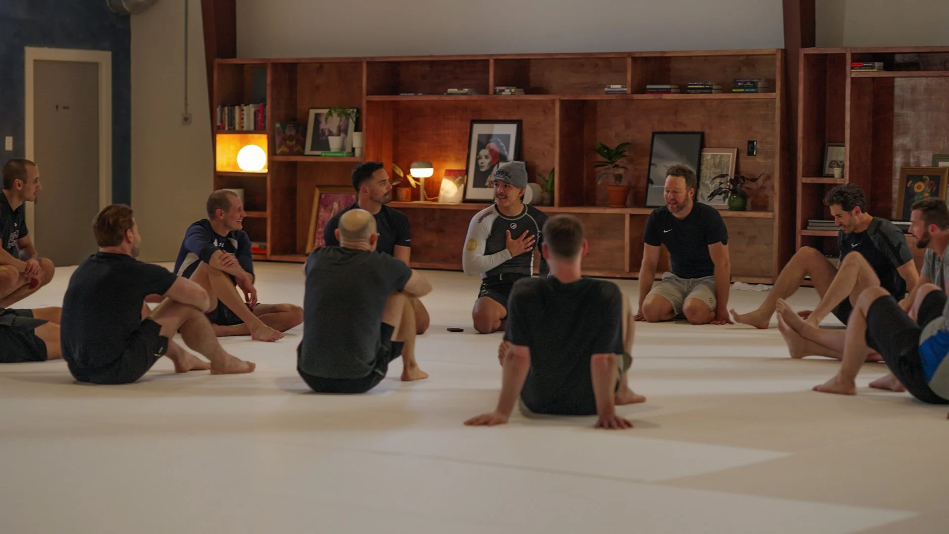 Group of people practicing yoga or stretching on the floor in a room with bookshelves and framed pictures on the wall, some people sitting cross-legged and others with legs extended.