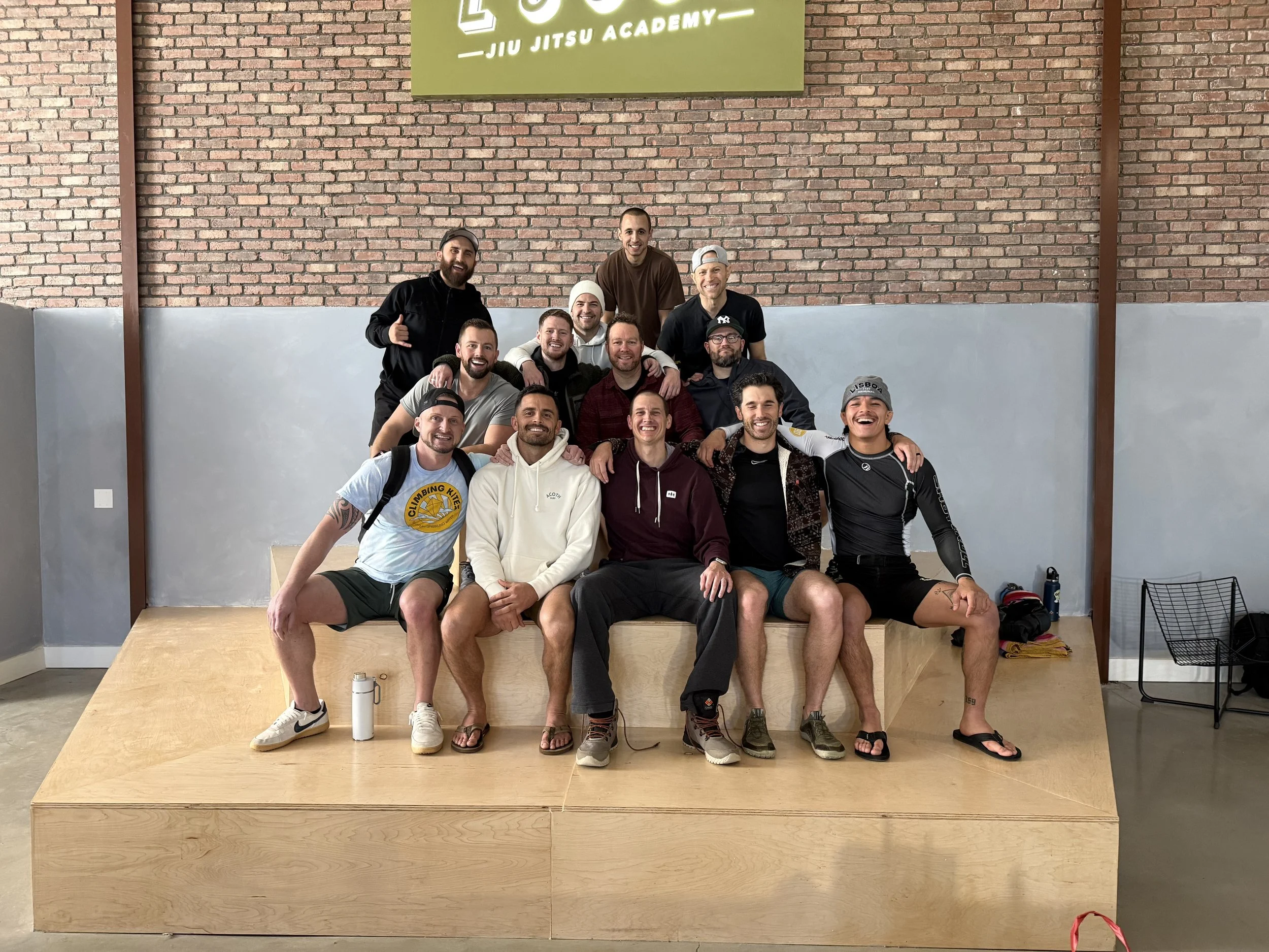 A group of fifteen men posing for a photo inside a gym or fitness center, standing and sitting on a wooden platform against a brick wall with a green sign that says 'Jiu Jitsu Academy'.