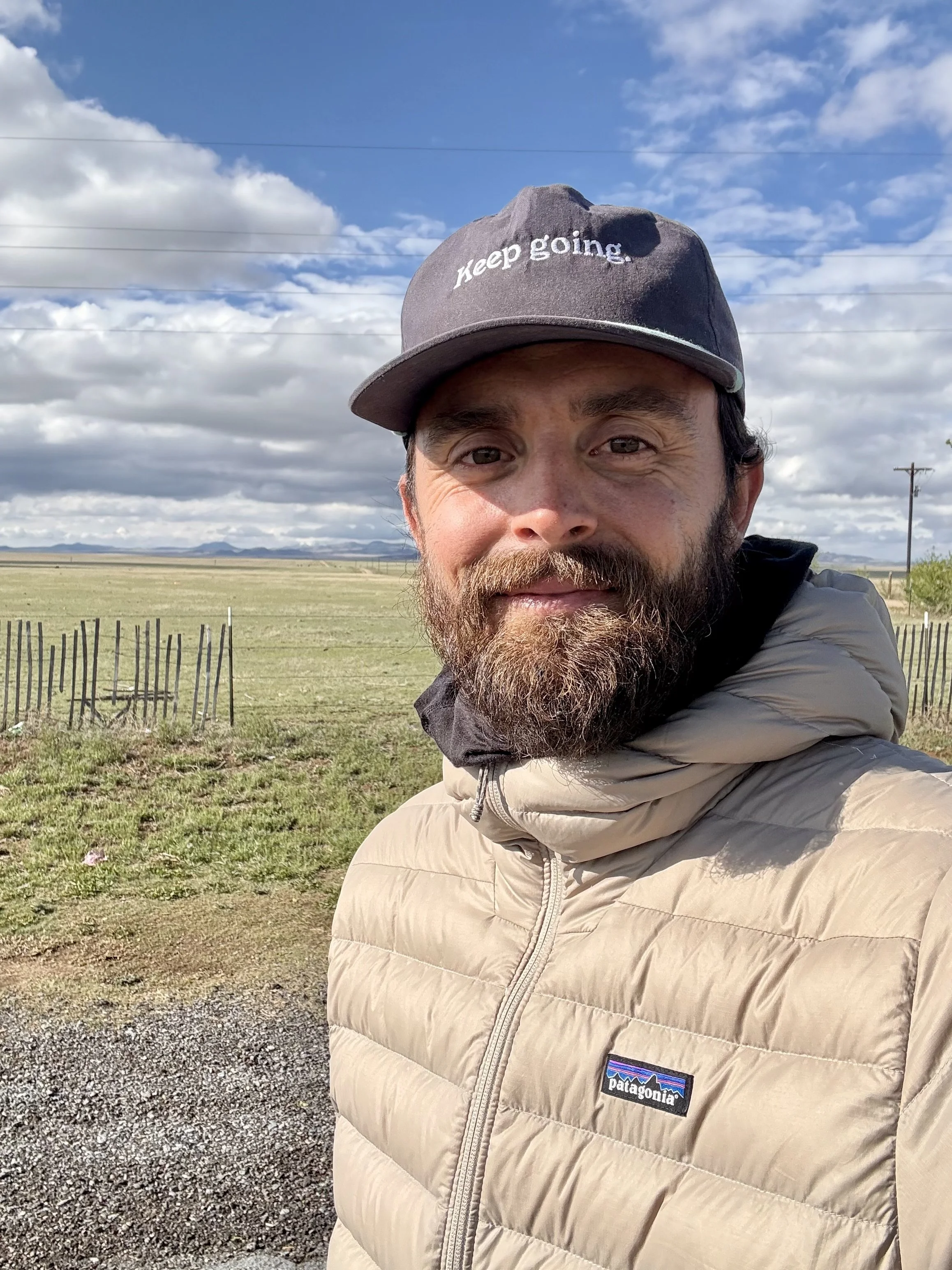 A man with a beard and mustache taking a selfie outdoors during the daytime. He is wearing a dark gray baseball cap that says 'keep going.' and a beige Patagonia puffer jacket. The background features a vast open field with a few small trees, a wooden fence, distant mountains, and a partly cloudy sky with blue and white clouds.