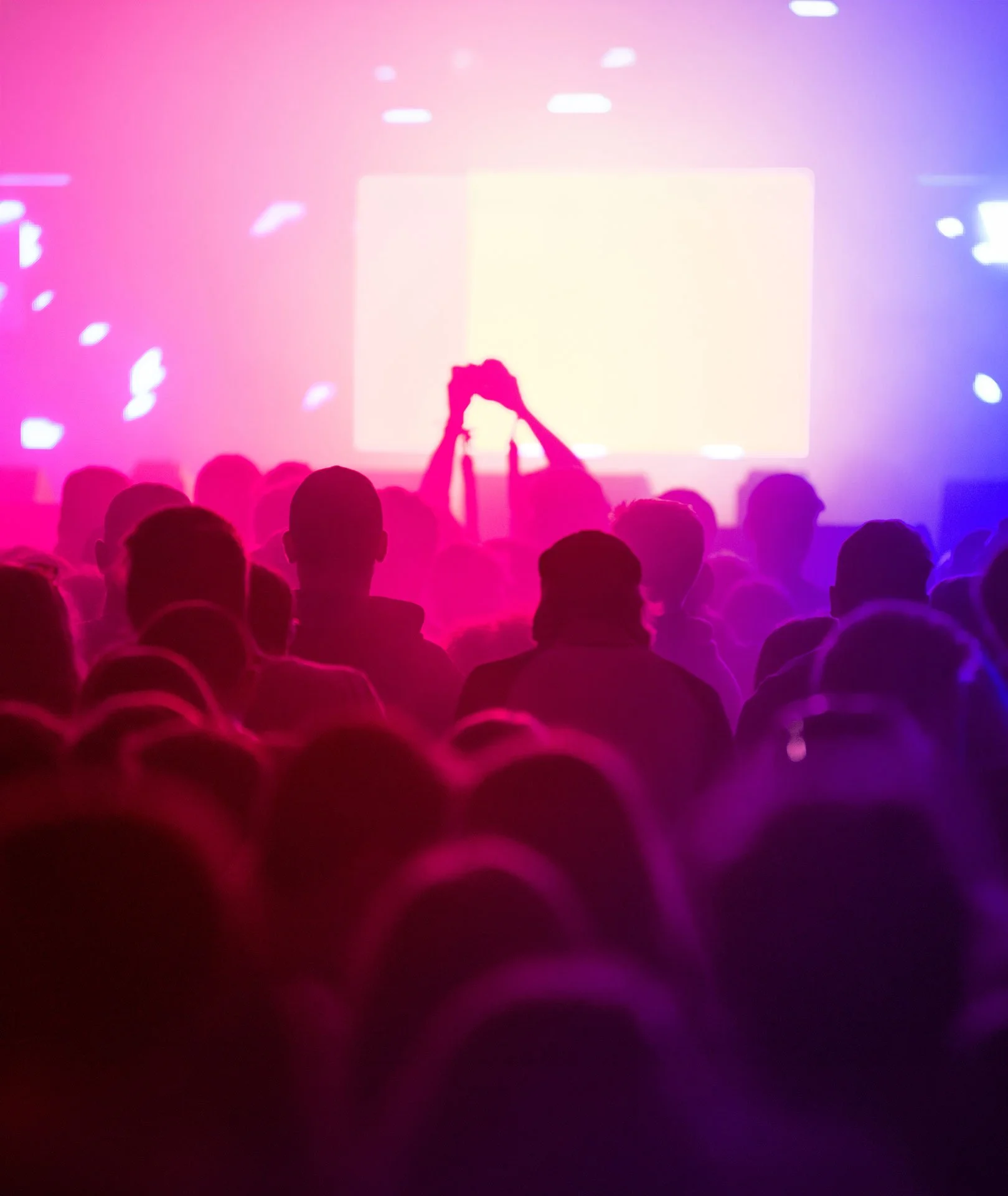 A crowd of people at an indoor event with a large lighted screen in the background, illuminated by pink and purple lighting.
