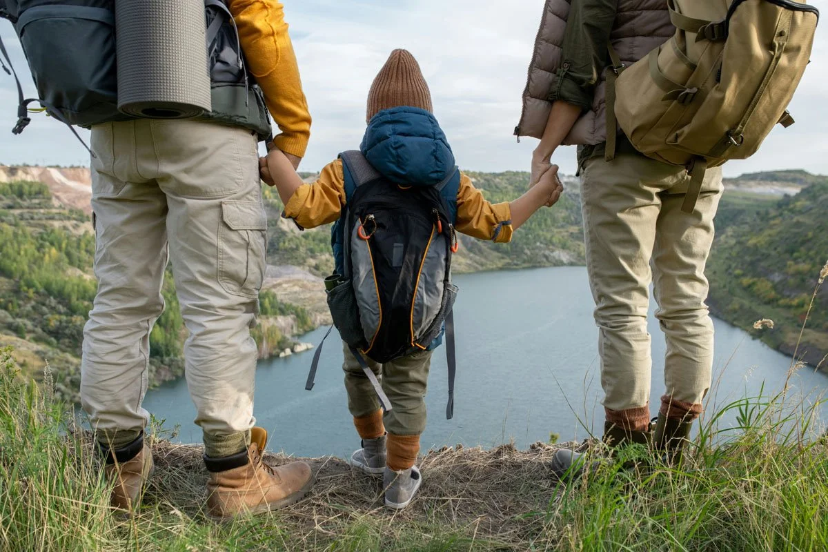 A family of three holding hands while standing on a hilltop overlooking a lake surrounded by green hills during daytime.