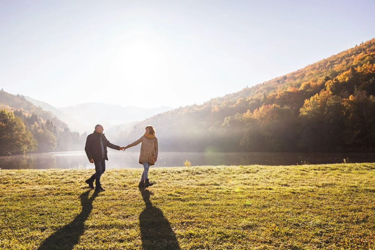 A couple holding hands and walking on grassy terrain near a lake, with mountains and trees in the background, during sunset.