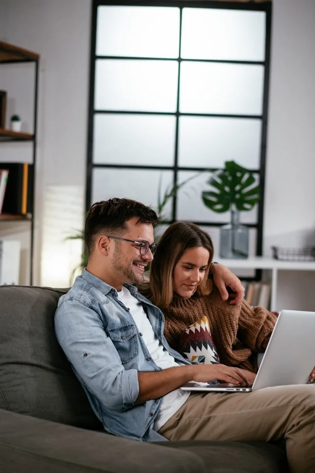 A young man and woman sitting on a gray sofa, looking at a laptop together in a modern, well-lit room with large window.