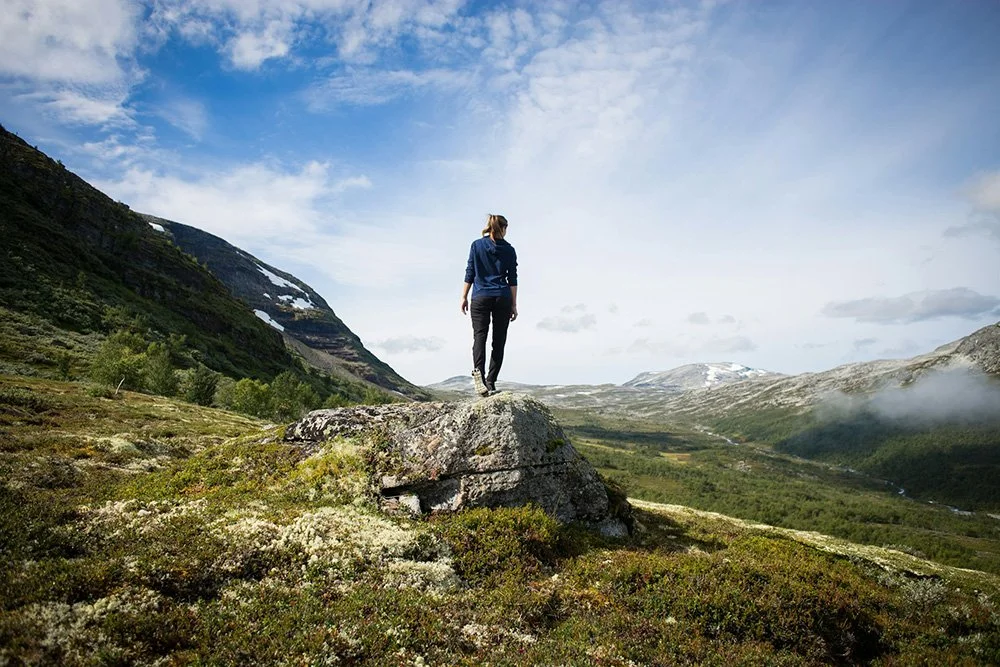 A woman standing on a large rock overlooking a lush green valley with mountains in the background and a partly cloudy sky.
