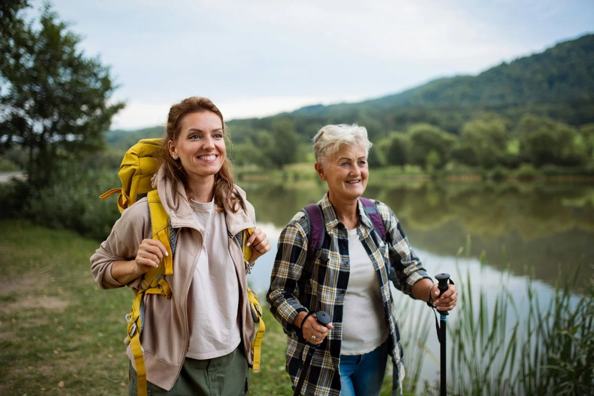 A mother and her adult daughter smiling and standing outdoors near a lake, with hills and trees in the background.