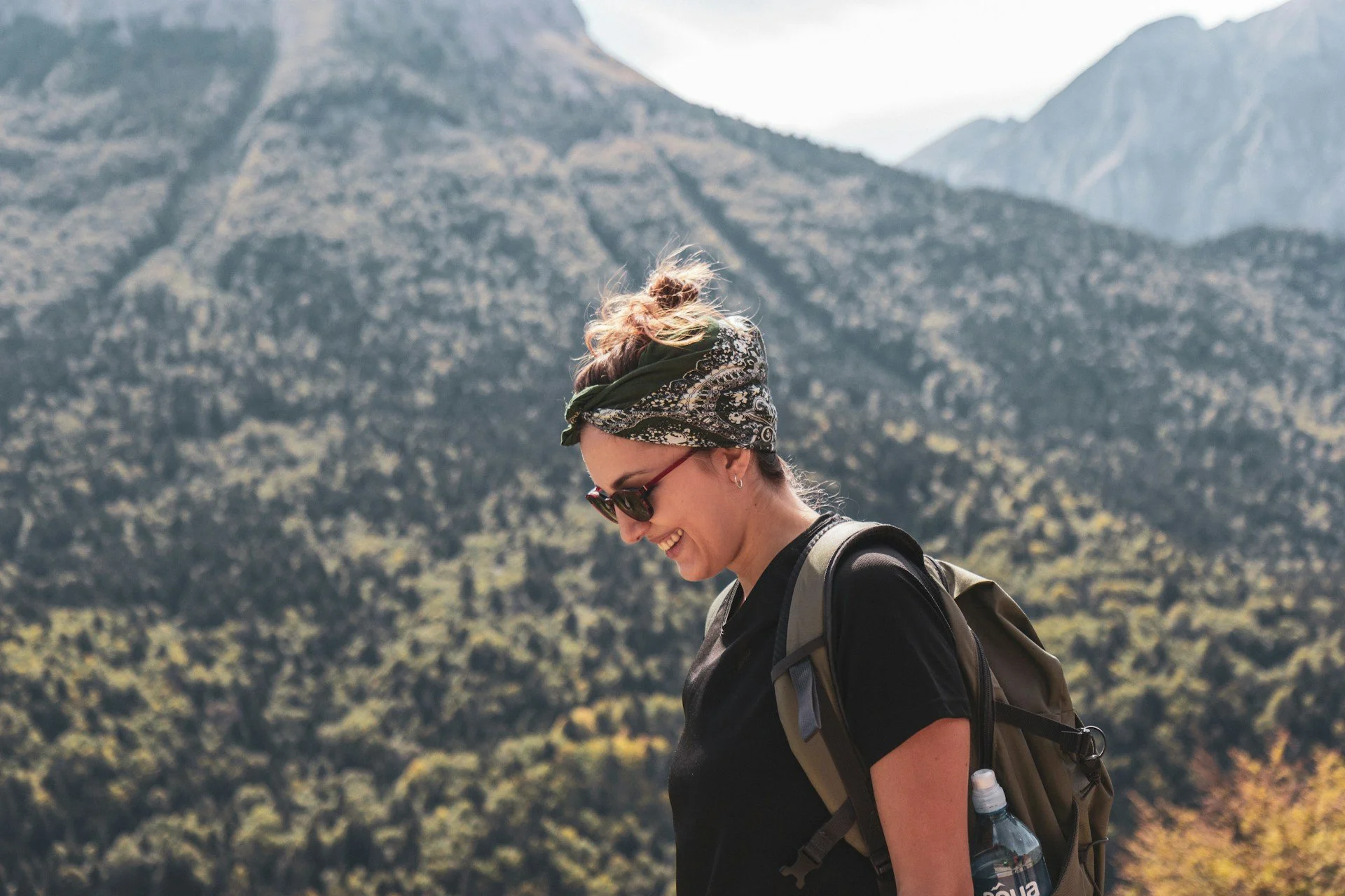 A woman hiking outdoors with mountains in the background, wearing sunglasses, a bandana, a black shirt, and a backpack.