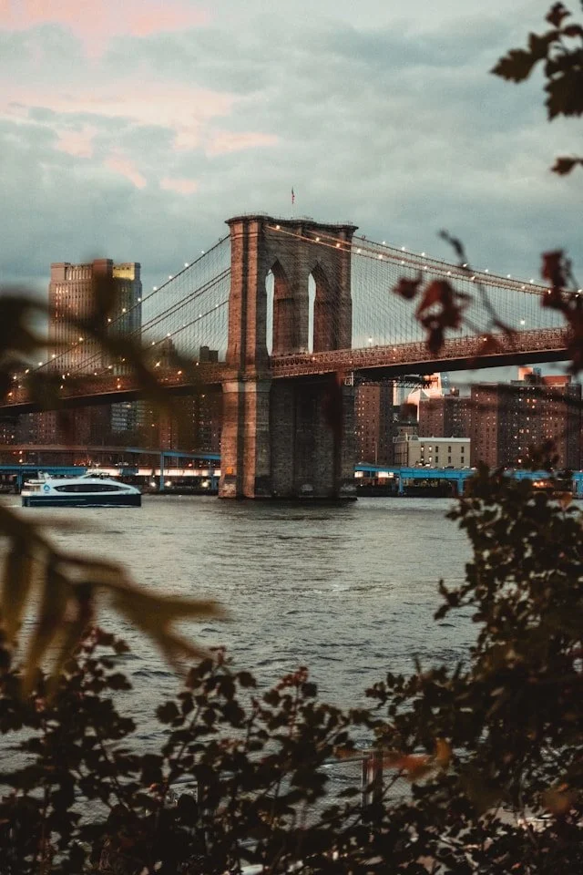 Brooklyn Bridge at dusk with city skyline and river in the foreground