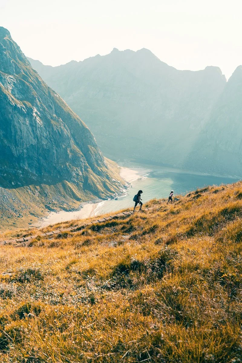 Two hikers walking along a mountain trail overlooking a scenic valley with mountains in the background and a river flowing through the valley.