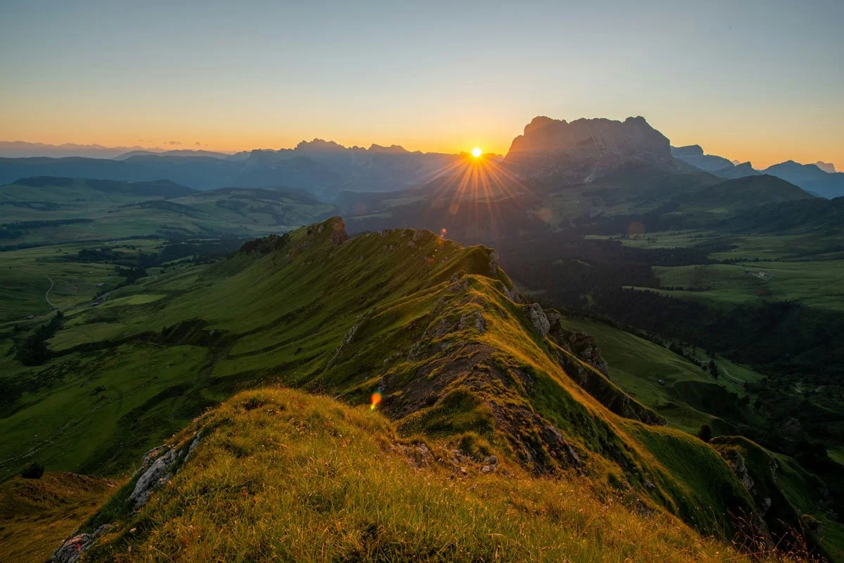 Sunset over green rolling hills and distant mountains, with a ridge in the foreground.