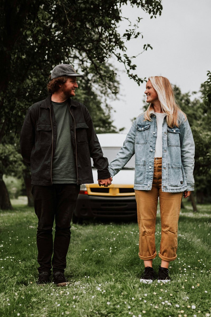 A couple holding hands and sharing a smile in a park with a vehicle in the background.