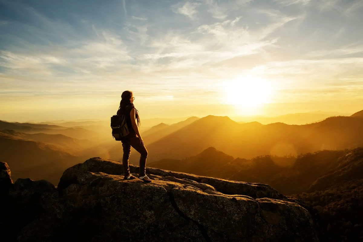 A person with a backpack standing on a rocky ledge overlooking mountains at sunrise.
