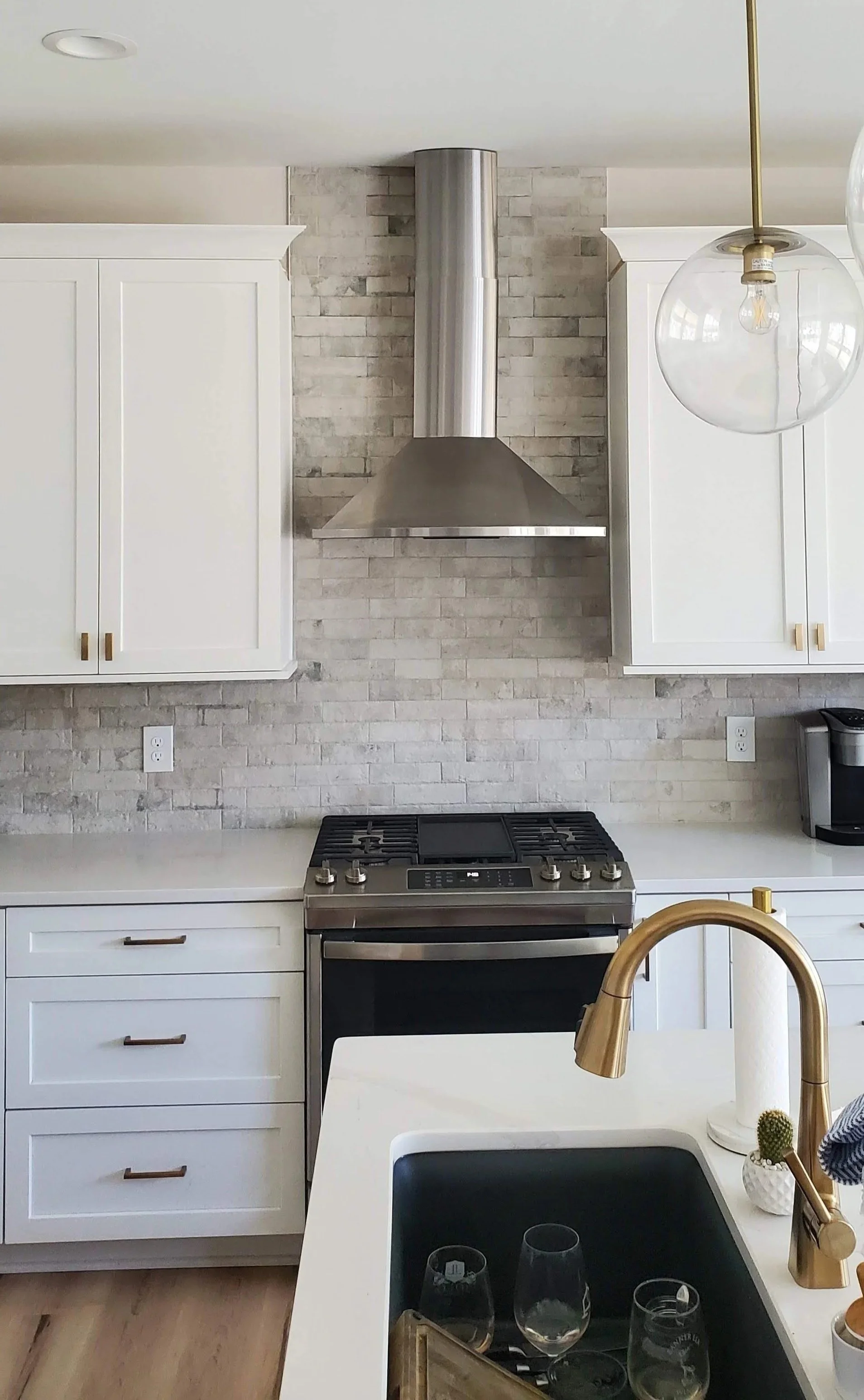 Modern kitchen with white cabinets, a stainless steel stove, brick backsplash, and a stainless steel range hood. A white kitchen island with a gold faucet and black sink is visible, with wine glasses and a tray. A hanging clear glass pendant light and a small cactus in a white pot are also included.