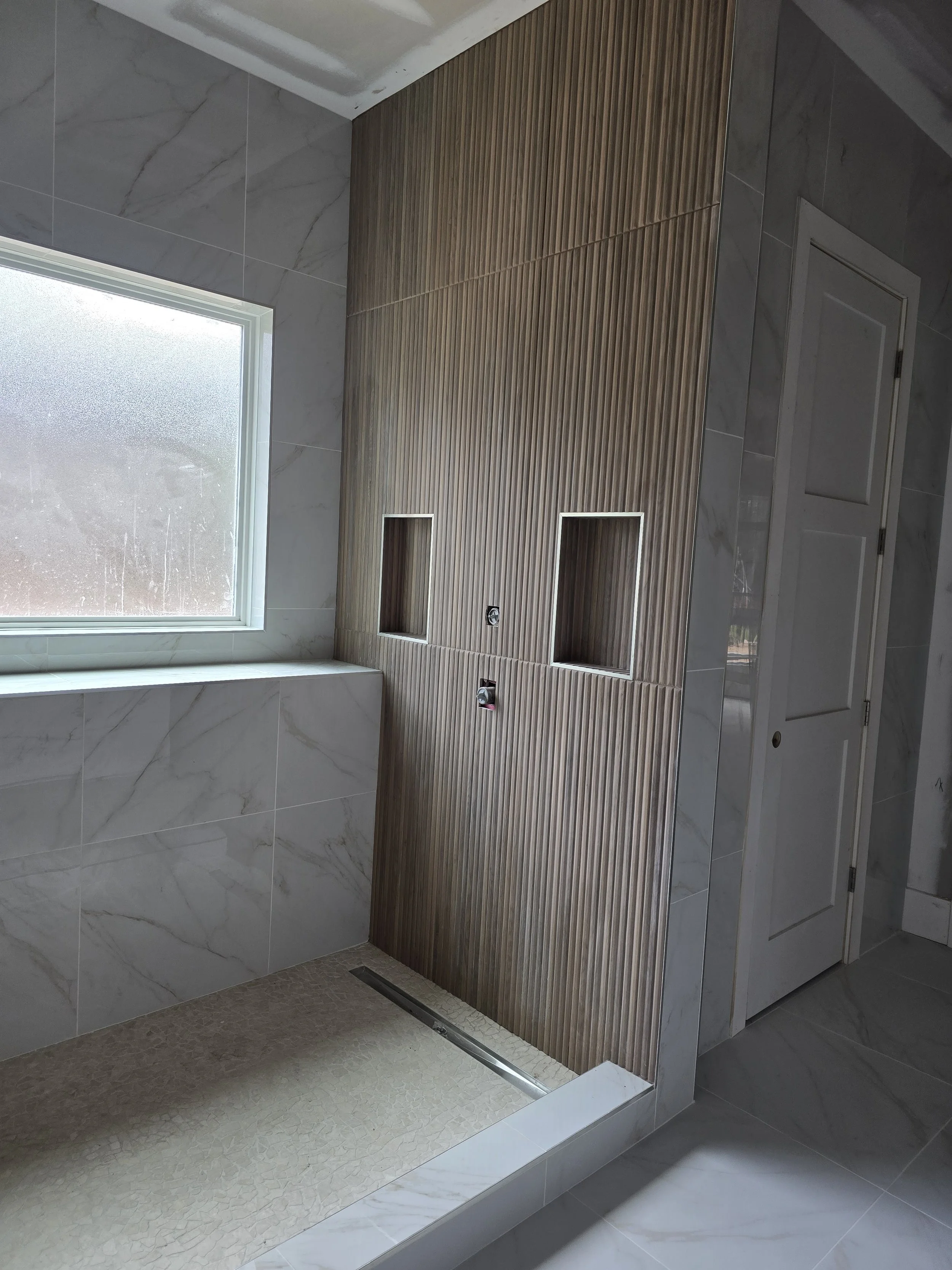 Bathroom with marble tiles, a frosted window, and a wooden accent wall with recessed shelves and plumbing fixtures.