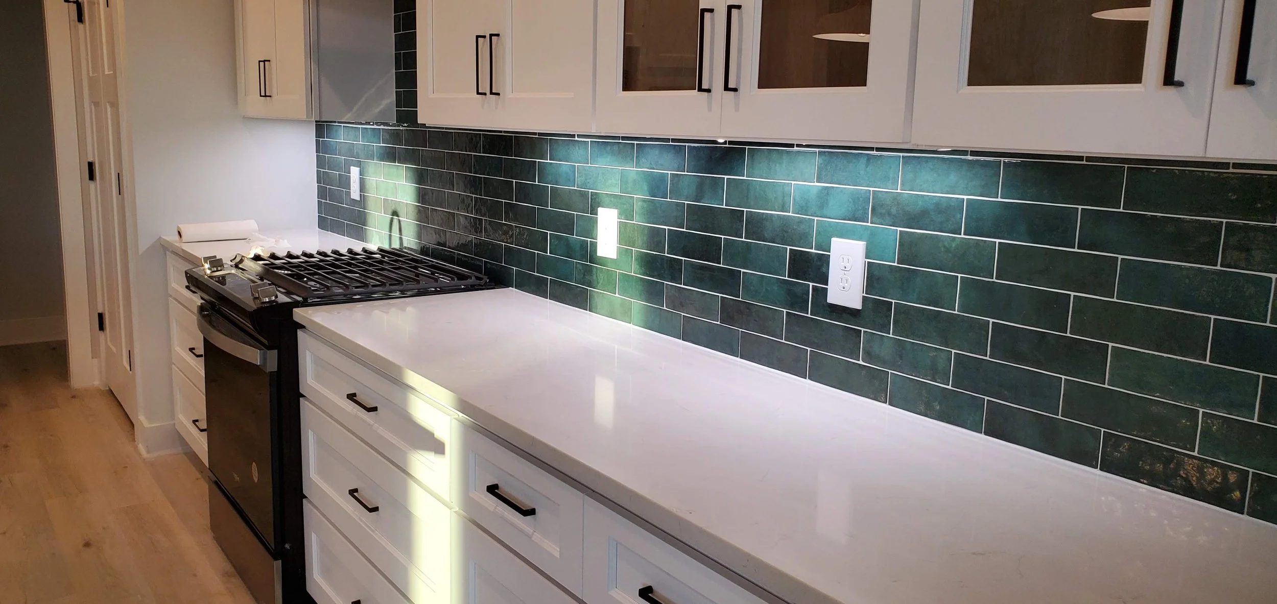 Modern kitchen with white cabinets, black handles, and a green subway tile backsplash. A black stove is installed on the white countertop, and there are electrical outlets on the backsplash. Wooden flooring is visible.