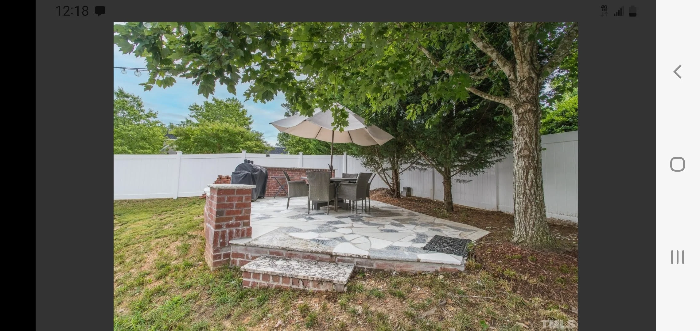 Backyard patio with a round table, six chairs, an umbrella, a brick barbecue, a white privacy fence, trees, string lights, and a stone-paved ground.