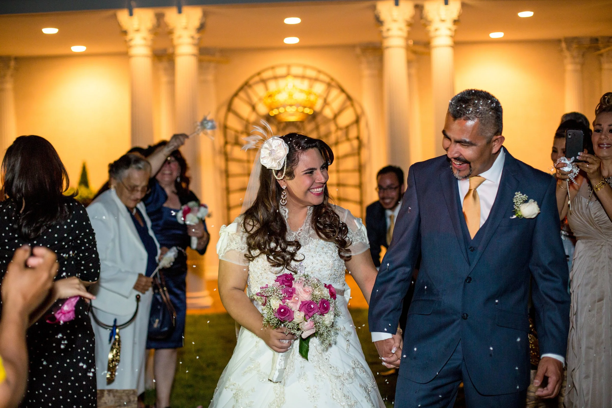Bride and groom smiling and holding hands at their wedding reception, surrounded by guests throwing confetti indoors with elegant columns and lighting.