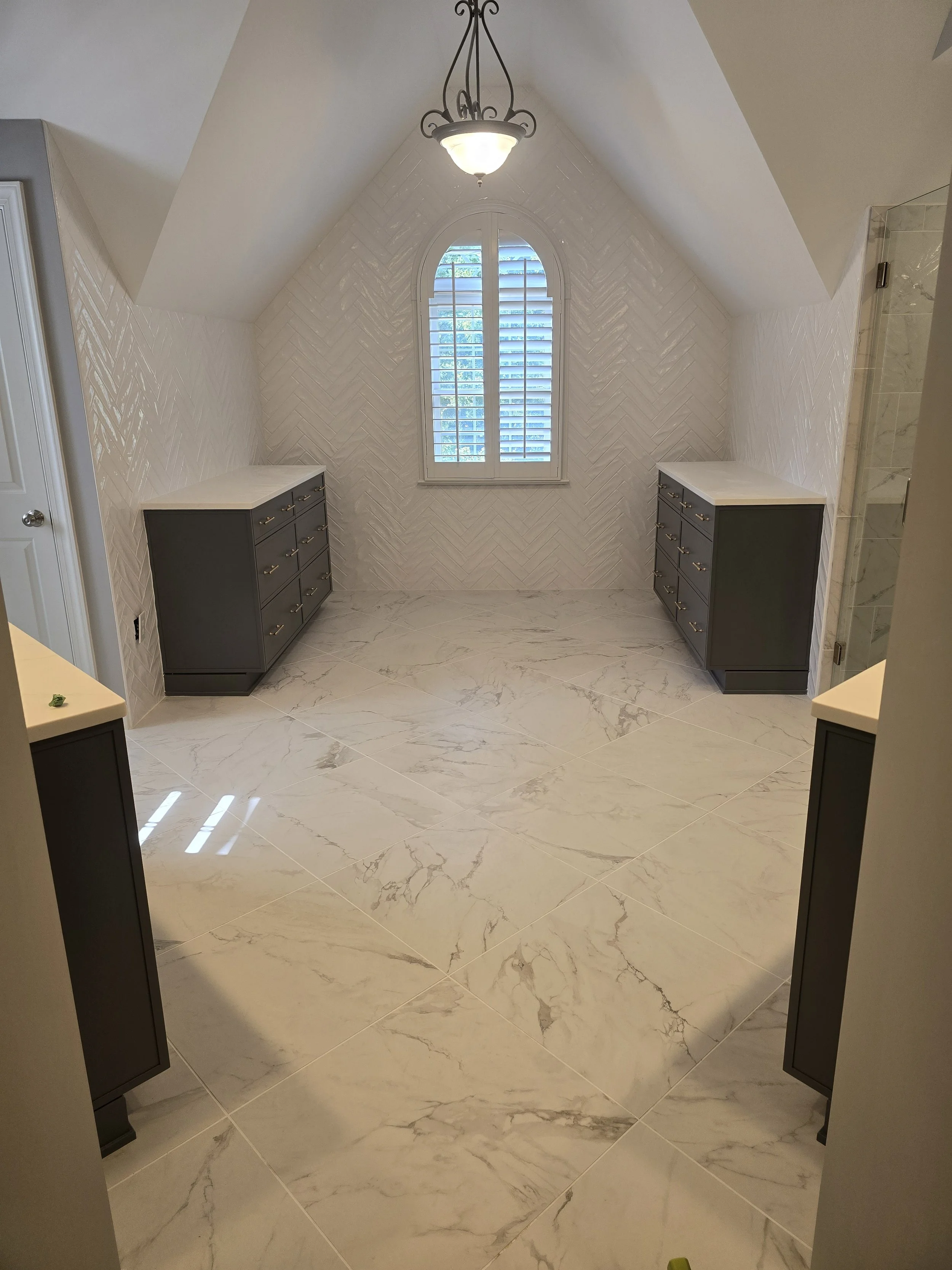 View of a bathroom with a large window with shutters, a chandelier, and two gray cabinets with white countertops on either side.