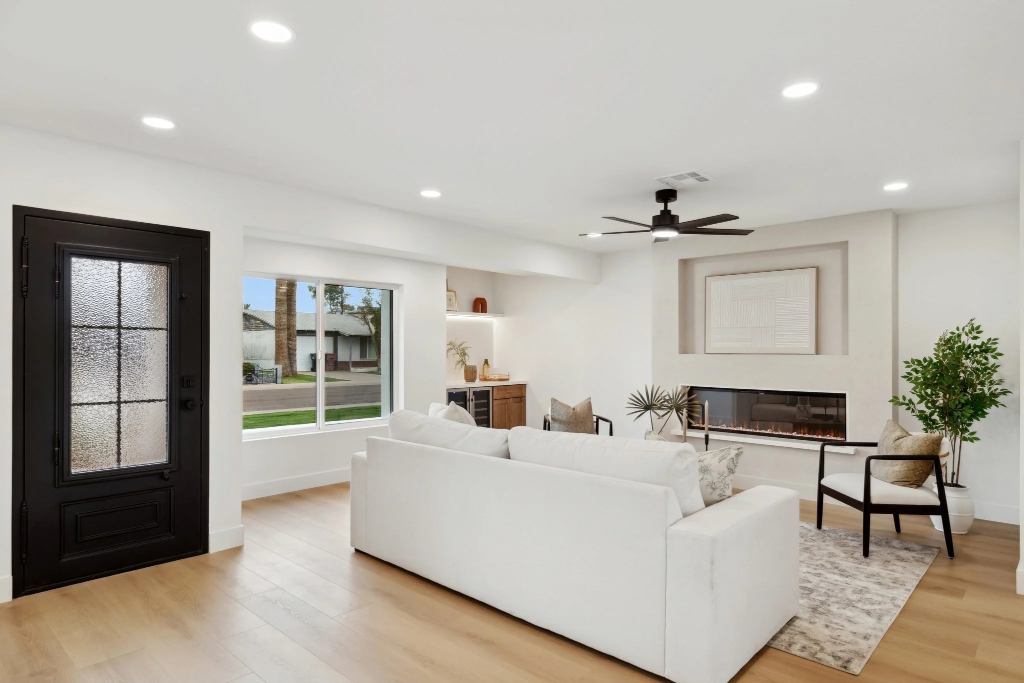 Living room with white sofa, chairs, wood floor, fireplace, black front door, large window, ceiling fan, wall art, and potted plant.
