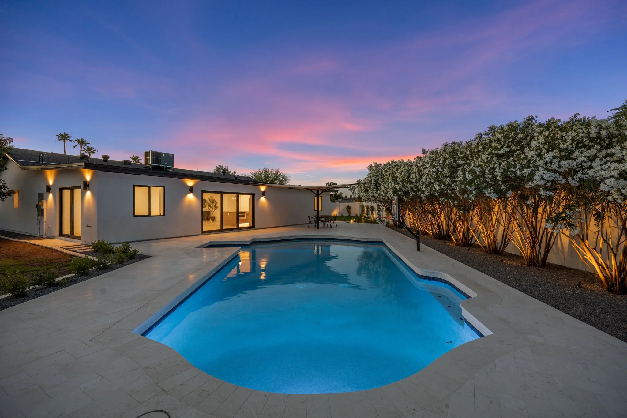 A modern house with large glass doors overlooking a backyard with a pool and landscaped garden, during sunset with colorful sky.