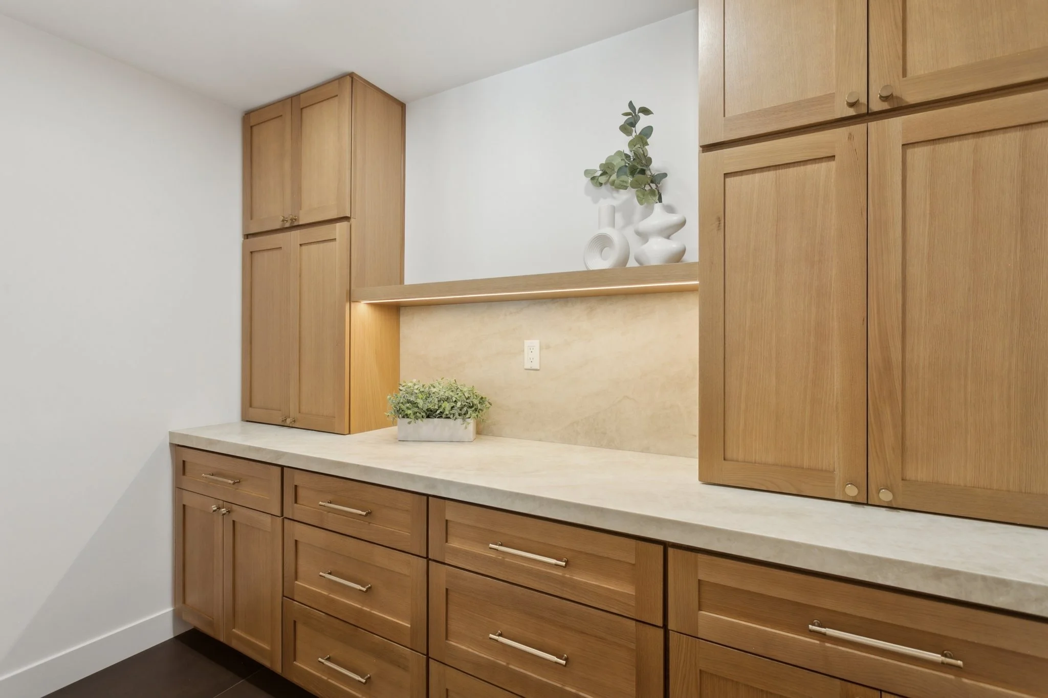 A modern kitchen countertop with wooden cabinets, white pottery vases with green foliage, a marble-like surface, and electrical outlets.