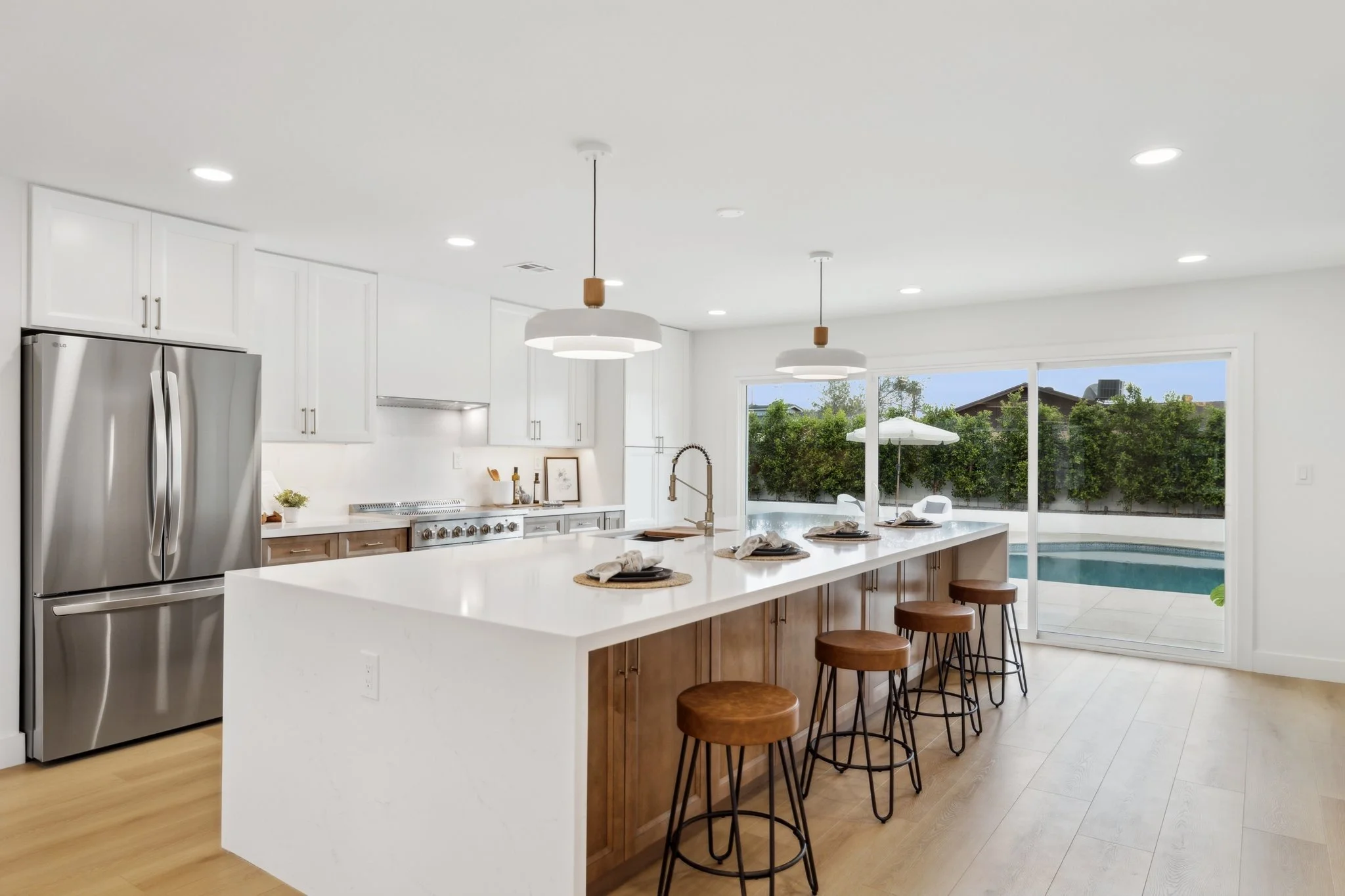 Modern kitchen with white cabinets, stainless steel refrigerator, island with bar stools, pendant lighting, and sliding glass doors leading to outdoor pool area.
