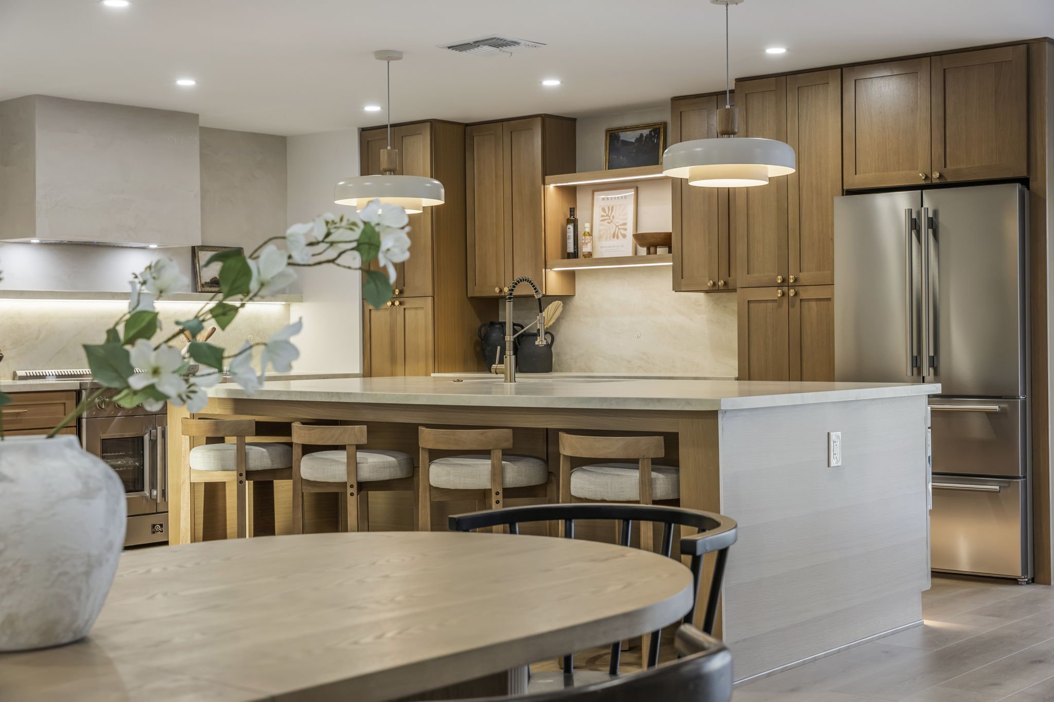 Modern kitchen with wood cabinetry and a large island with bar stools, pendant lighting, and a vase with white flowers in the foreground.