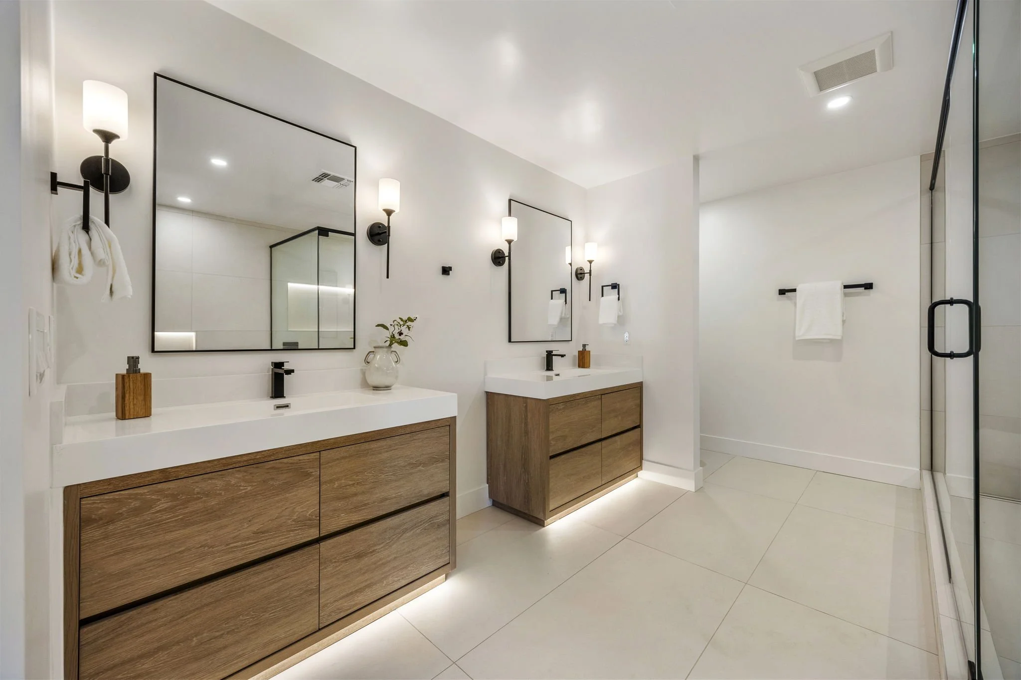 Modern bathroom with white walls and ceiling, black-framed mirrors above wooden vanities, black wall sconces with white shades, and a glass shower enclosure with a black handle. Decor includes a vase with greenery and a soap dispenser.