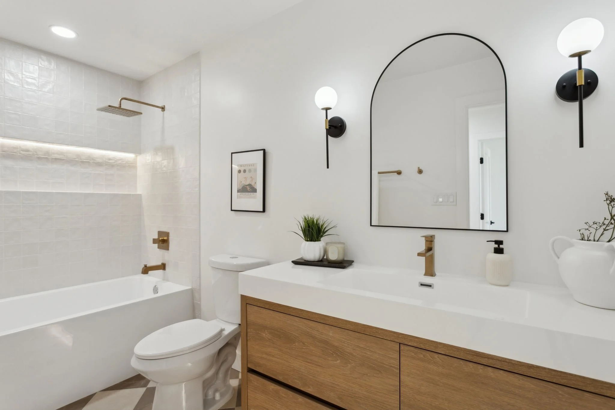 Modern bathroom with white walls, a large mirror, black and gold wall sconces, and a white vanity with wooden drawers. There is a bathtub with a gold faucet and a shower with a gold rain showerhead. Decor includes a framed art print, a potted plant, 