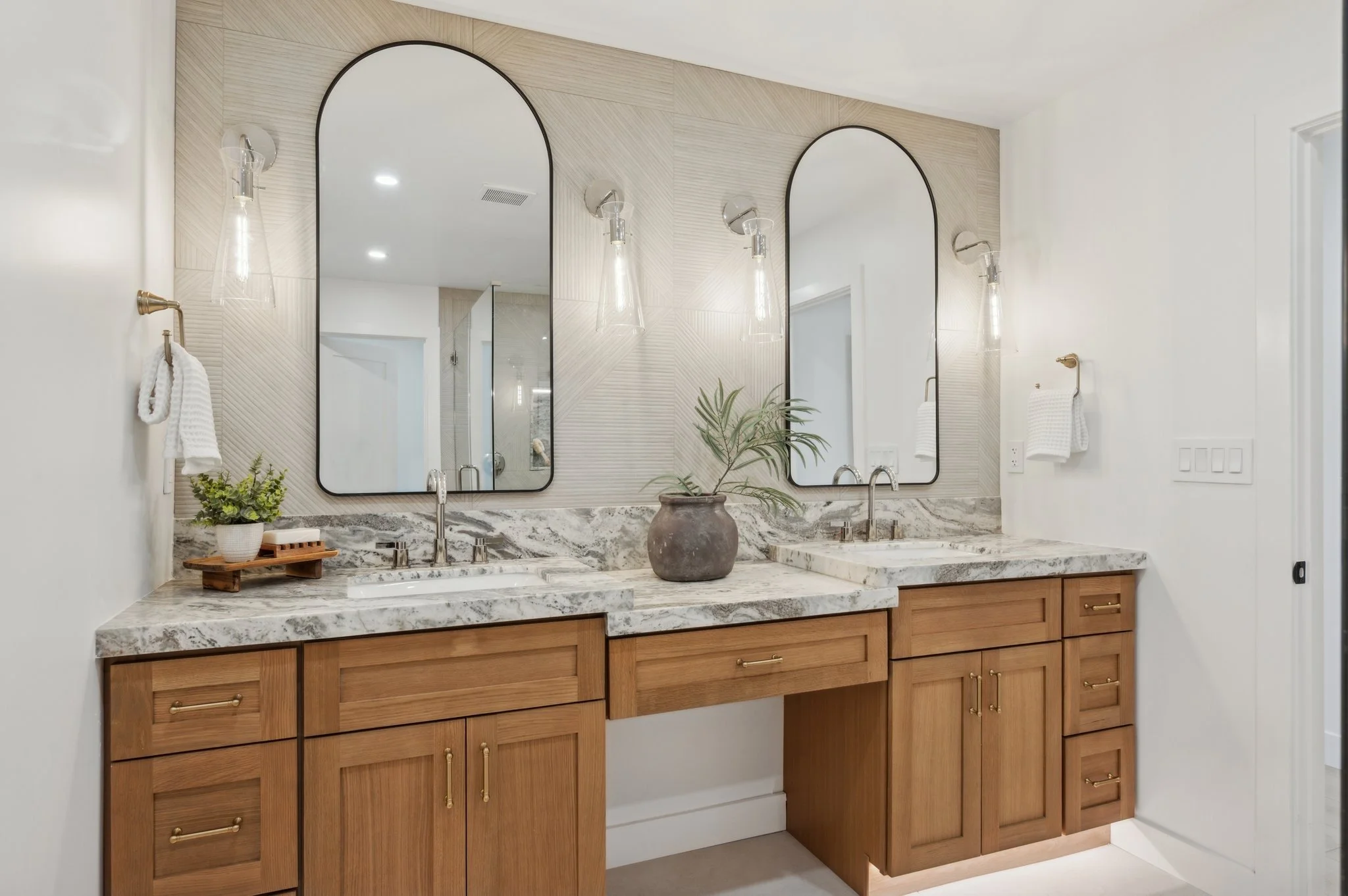 Bathroom vanity with two large mirrors, wooden cabinets, a marble countertop, two sinks, and two wall-mounted light fixtures. Decor includes a small potted plant and a tray with toiletries.