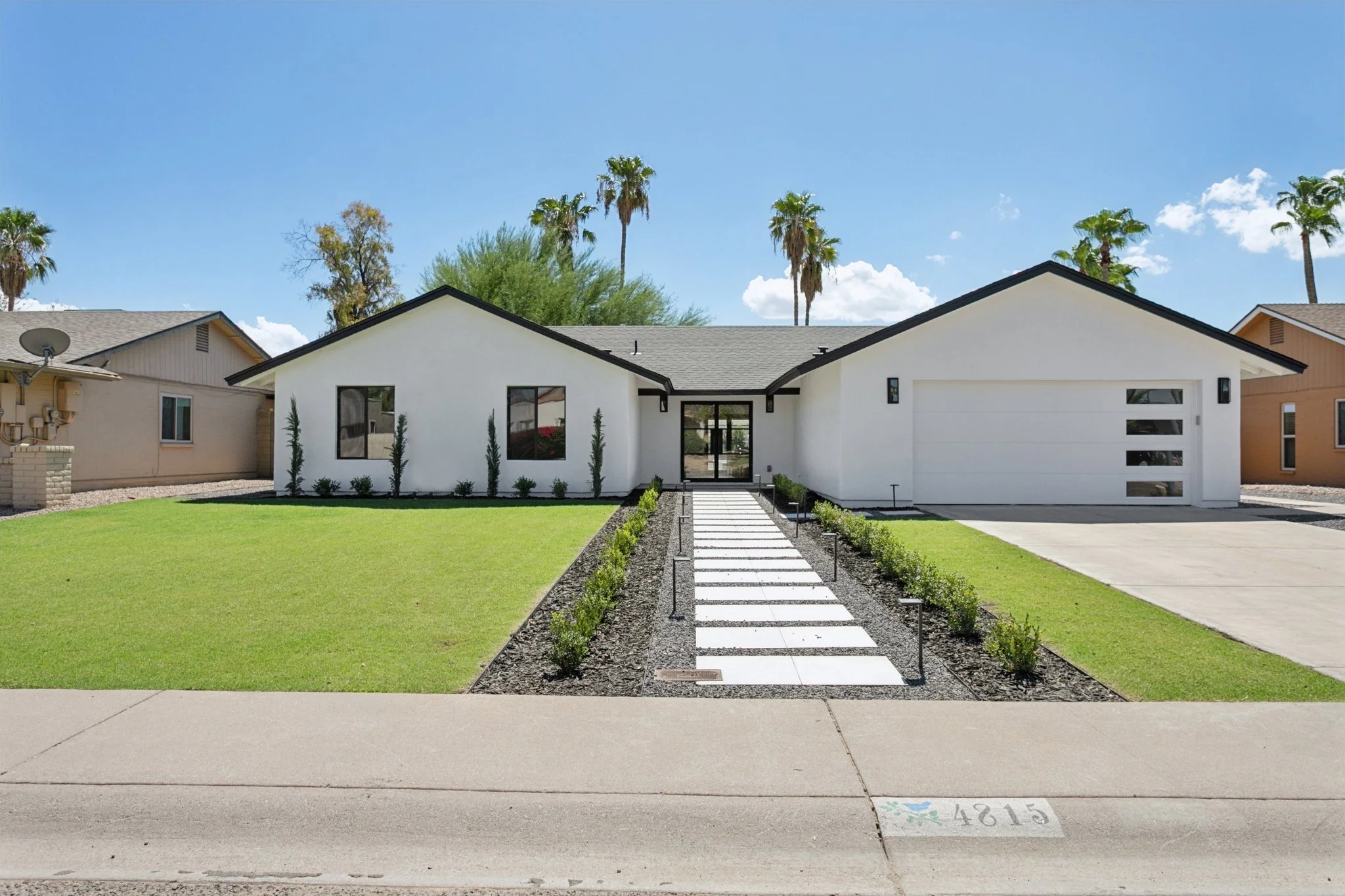Modern white house with a gray roof, front yard with a pathway, green lawn, and palm trees in the background under a blue sky.