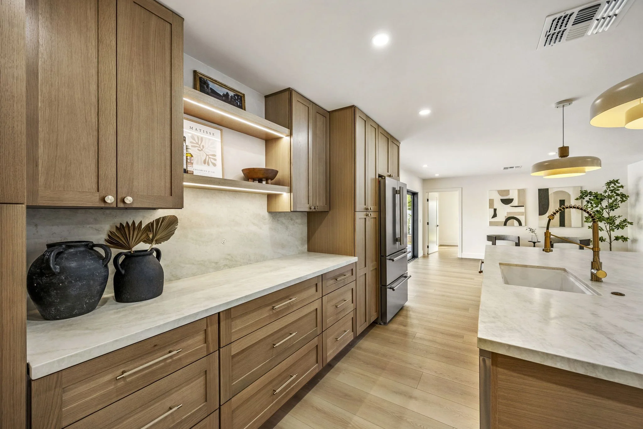 A modern kitchen with wooden cabinets and white marble countertops. The kitchen features a stainless steel refrigerator, a sink with a gold faucet, decorative vases, and open shelves with art and decorative items. The dining area in the background ha