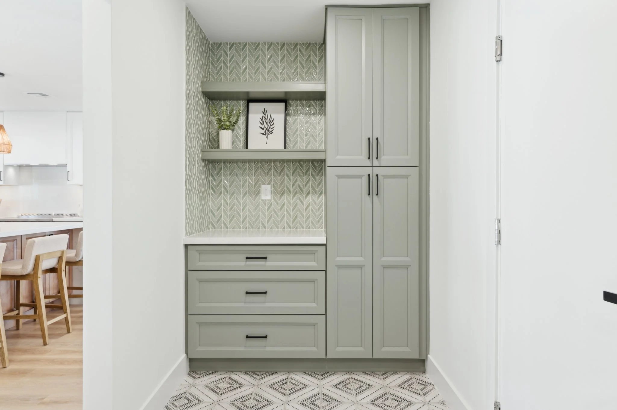 Interior of a kitchen with light gray cabinets, a patterned backsplash, and a built-in desk area with drawers and shelving, with a decorative plant and framed artwork on the shelf.
