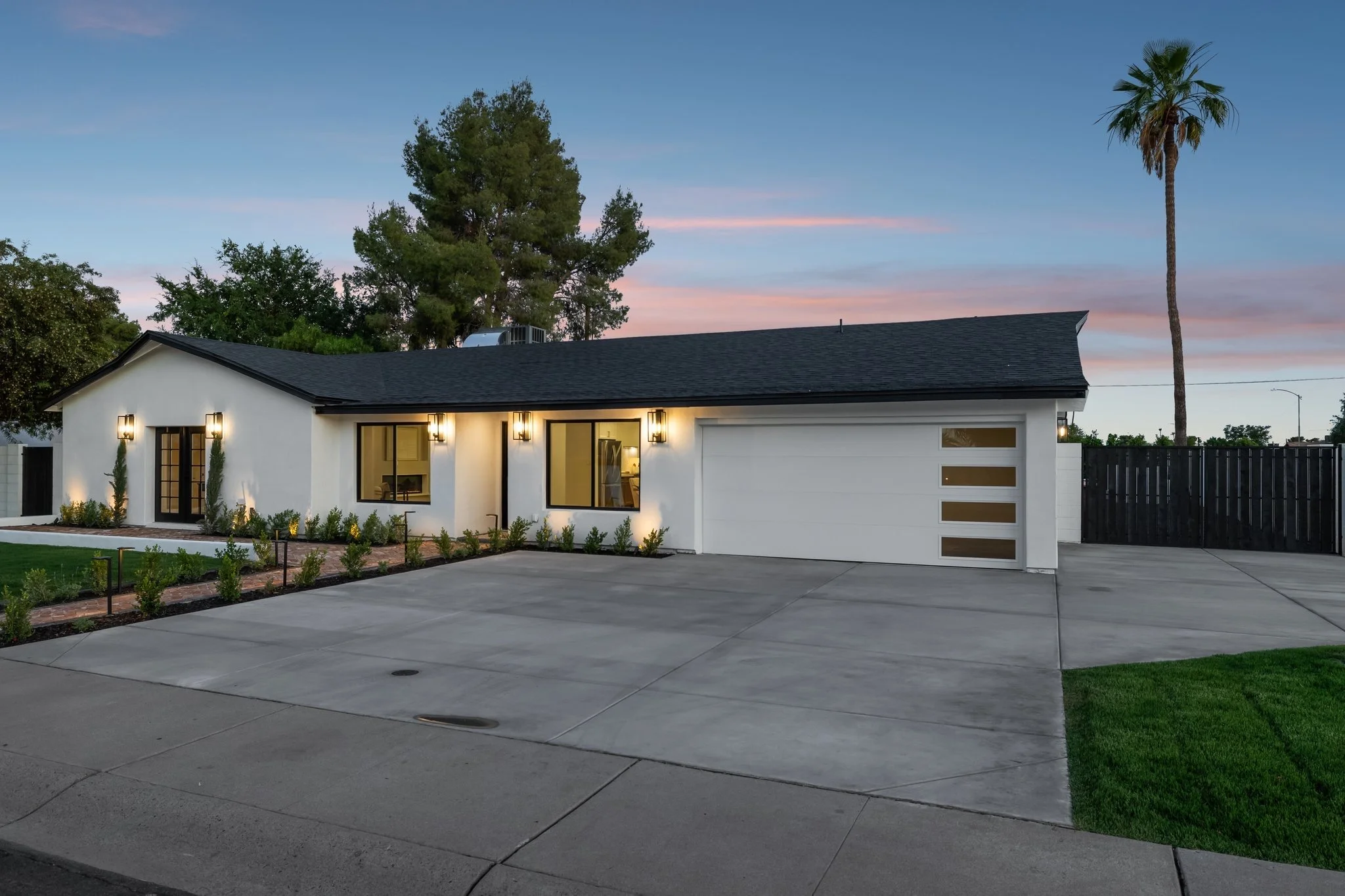 A modern white house with black window frames, outdoor lighting, a driveway, green landscaping, tall trees including a palm tree, and a pink and blue sky at dusk.