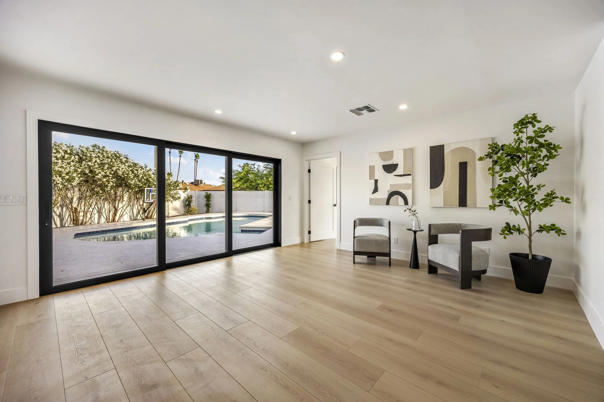 Empty living room with large sliding glass door showing backyard with pool and trees, white walls, wood flooring, two modern chairs, abstract art on wall, and a potted plant.