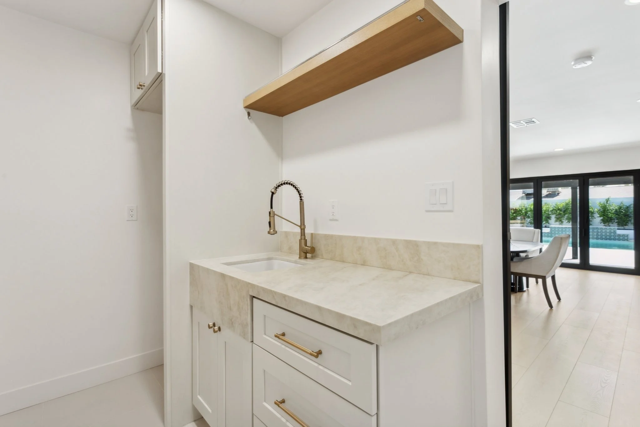 Interior view of a kitchen area with a light-colored marble countertop, white cabinets with gold hardware, a gold faucet, and a wood floating shelf above the sink. In the background, through a sliding glass door, there is a patio with greenery and ou
