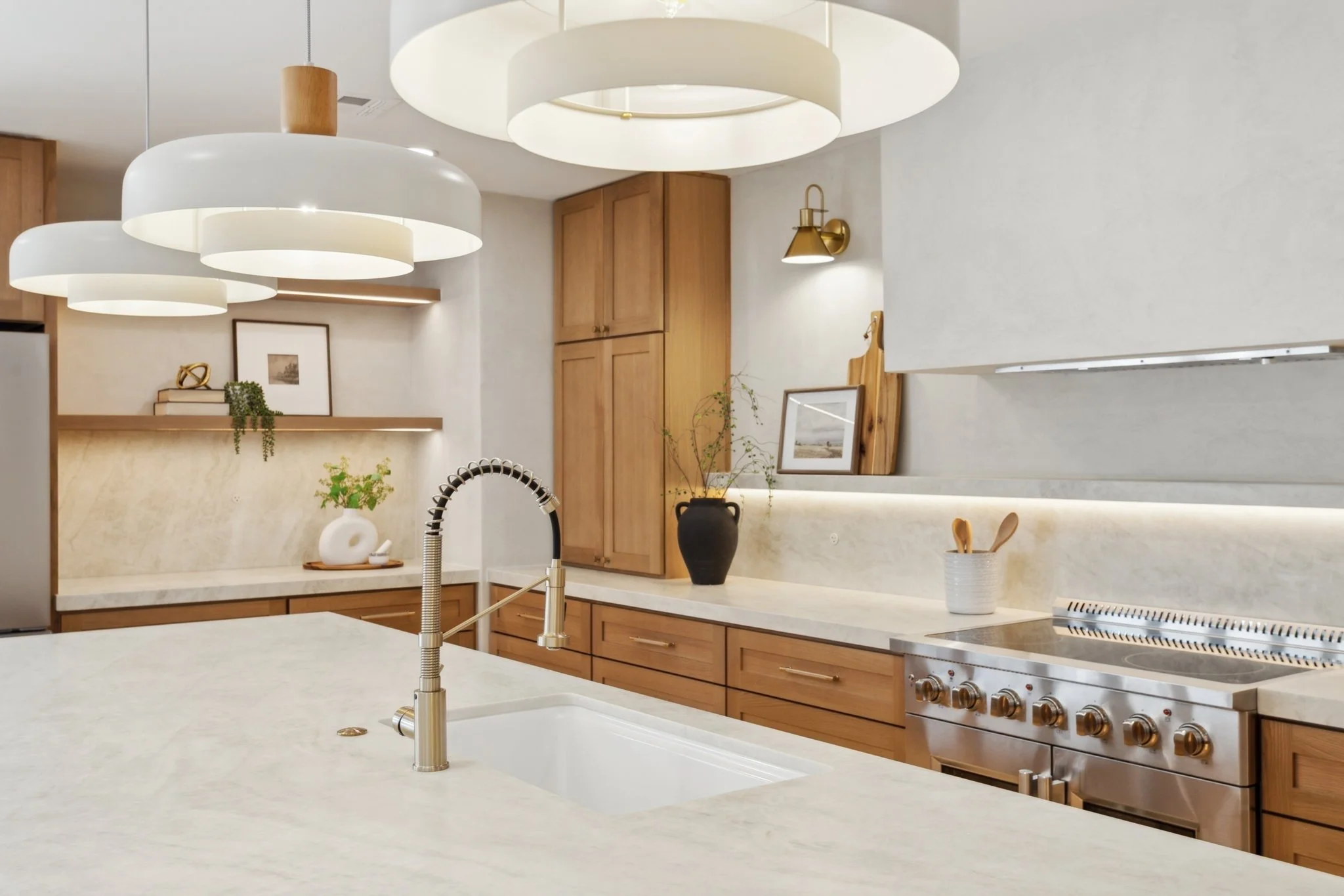 Modern kitchen with wooden cabinets, white marble countertops, and a stainless steel stove. Decorative items and plants are on the counter. Large white pendant lights hang from the ceiling.