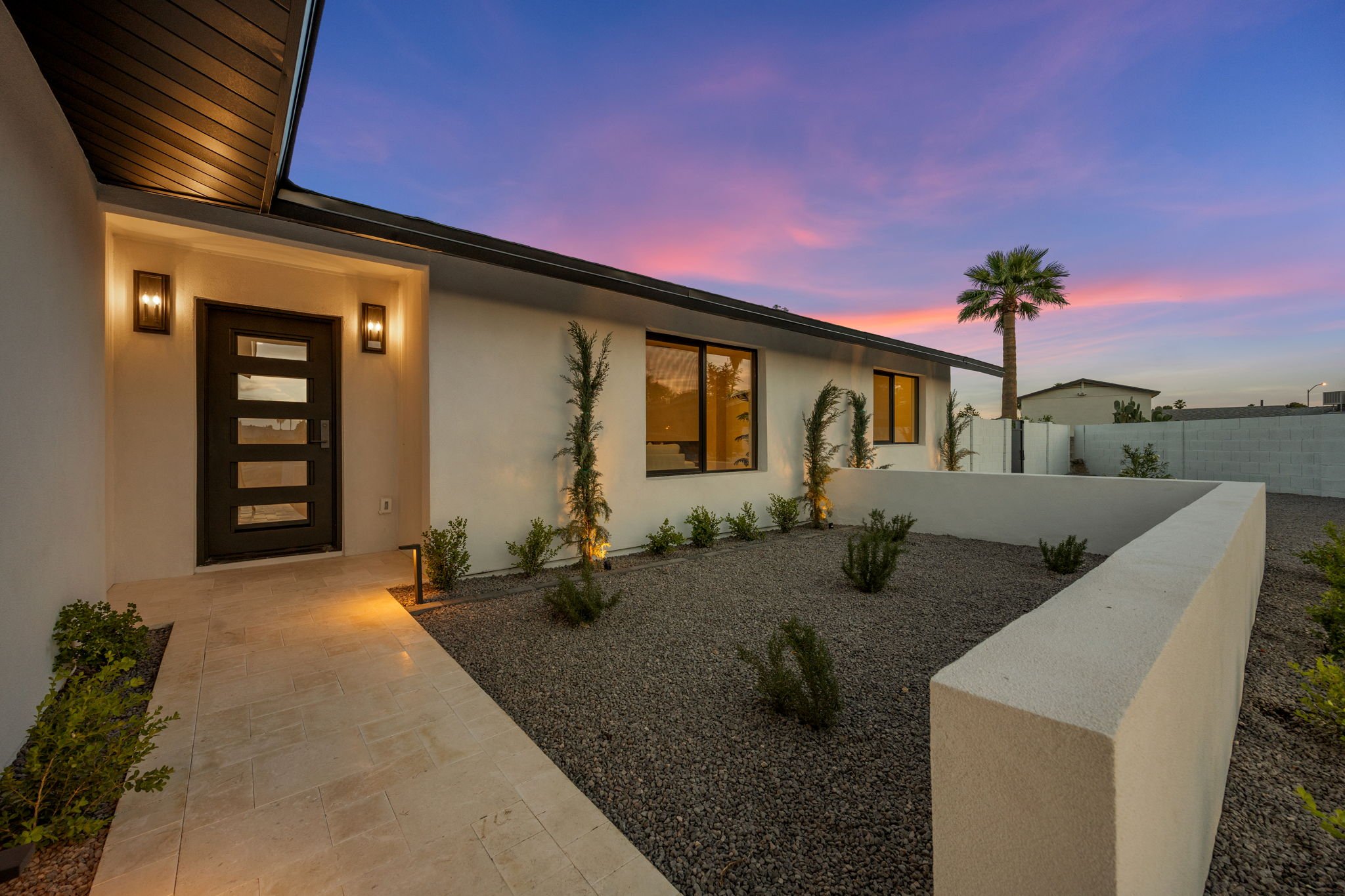 Modern house exterior at dusk with a sunset sky, featuring a black front door, large windows, palm tree, desert plants, and outdoor lighting.