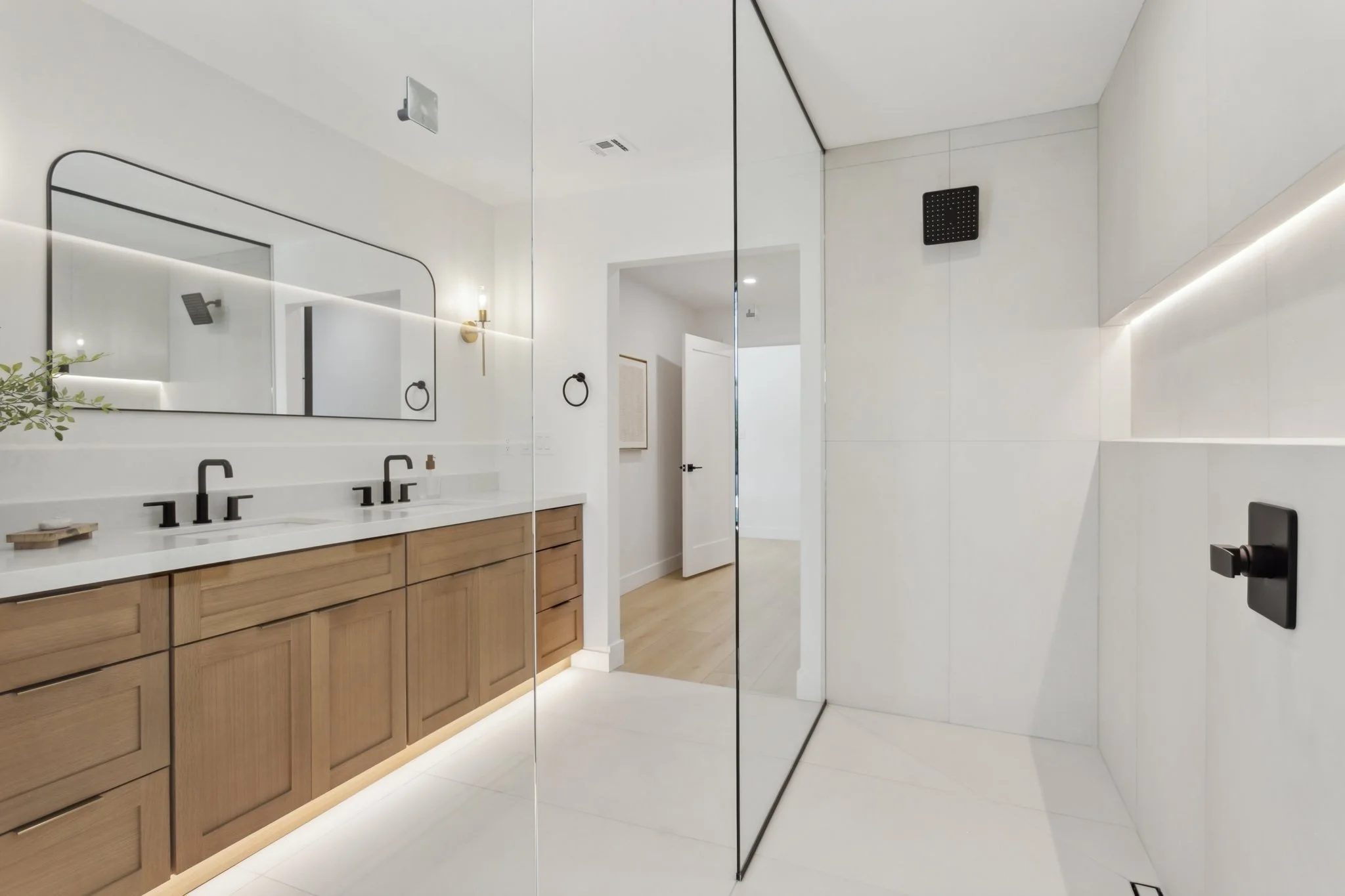 Modern bathroom with double sinks, wooden vanity, large mirror, black fixtures, white walls, and a walk-in shower with a black shower head and controls.