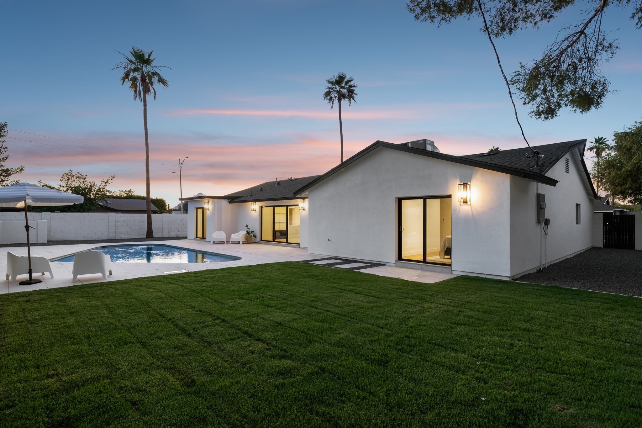 Contemporary backyard with a white house, a swimming pool, green lawn, patio furniture, and tall palm trees at sunset.