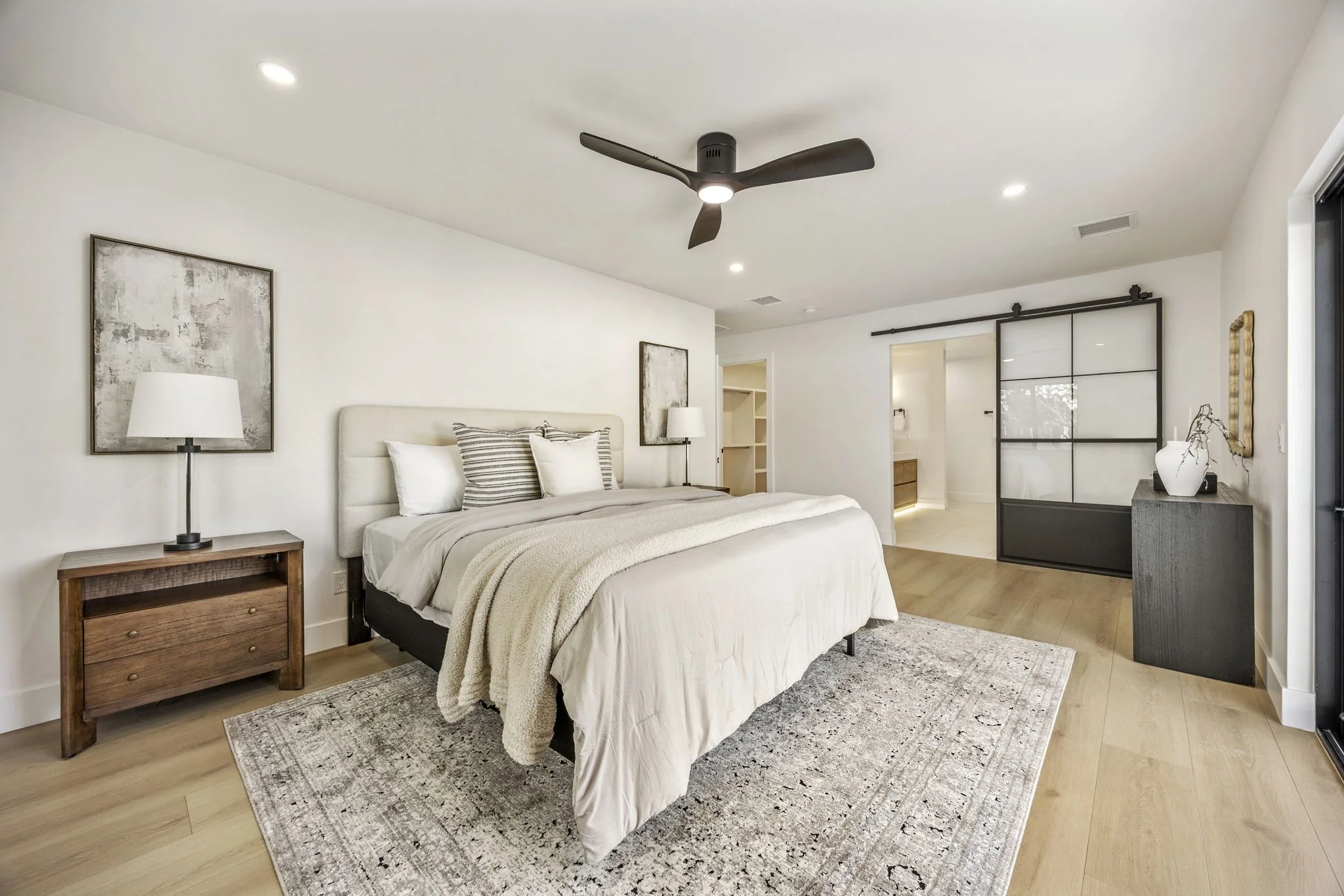 A modern bedroom with a queen-sized bed, white bedding, striped pillows, and a beige headboard. There are two abstract paintings on the white walls, two bedside tables with lamps, a gray side table with decorative items, and a ceiling fan with a ligh