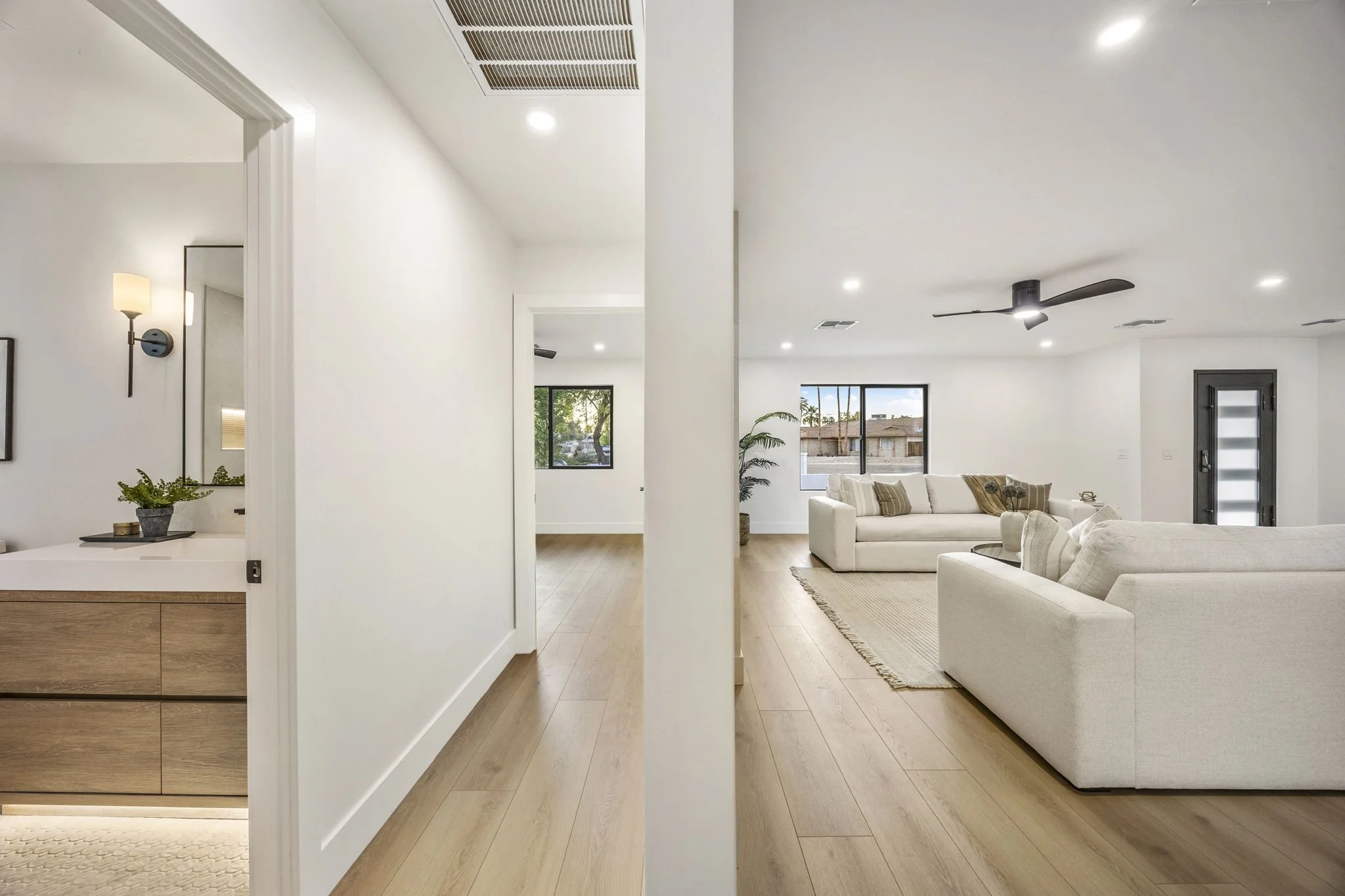 Open plan living room with white sofas, wooden flooring, large windows, ceiling fan, and a doorway with frosted glass.
