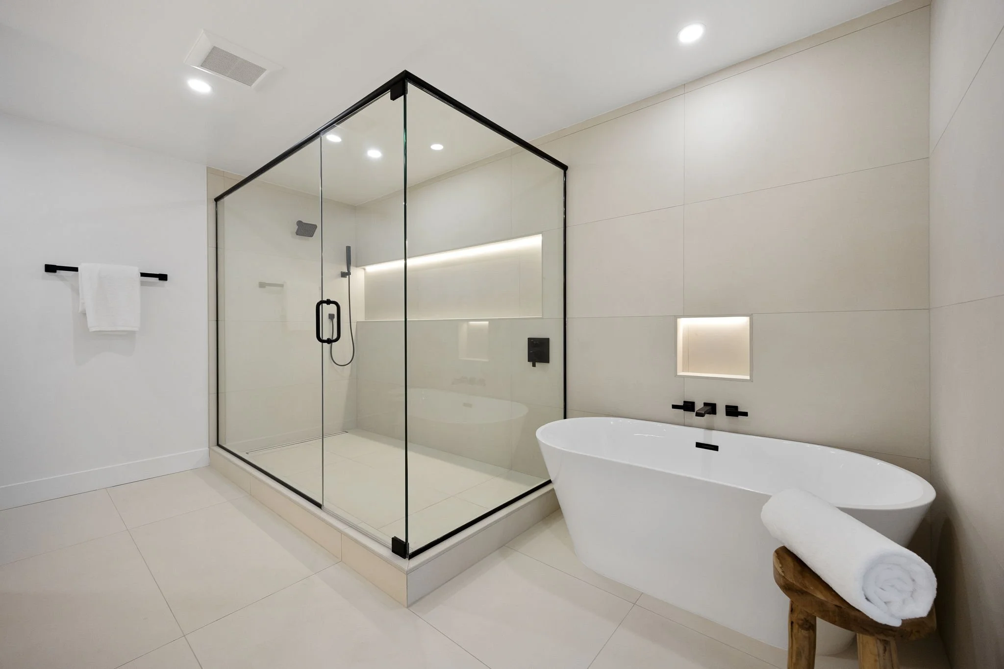 Modern bathroom with walk-in shower glass enclosure, black fixtures, white bathtub, and rolled white towels on a wooden stool.