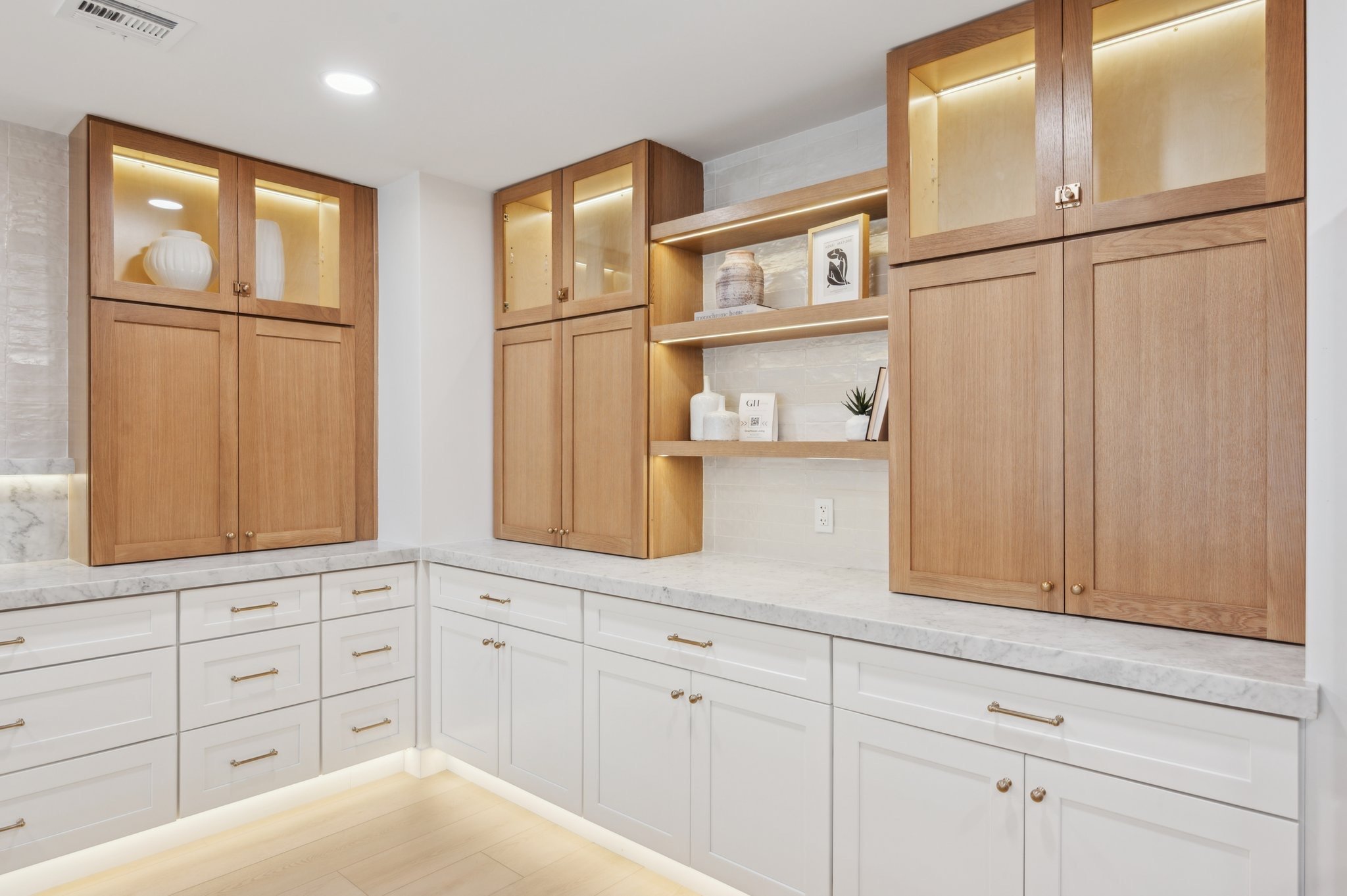 Kitchen cabinets with white drawers and gold handles, upper wooden cabinets with glass doors, decorative vases and framed art on open shelves, marble countertop, LED lighting beneath cabinets, cream-colored tile backsplash, ceiling light, air vent, e