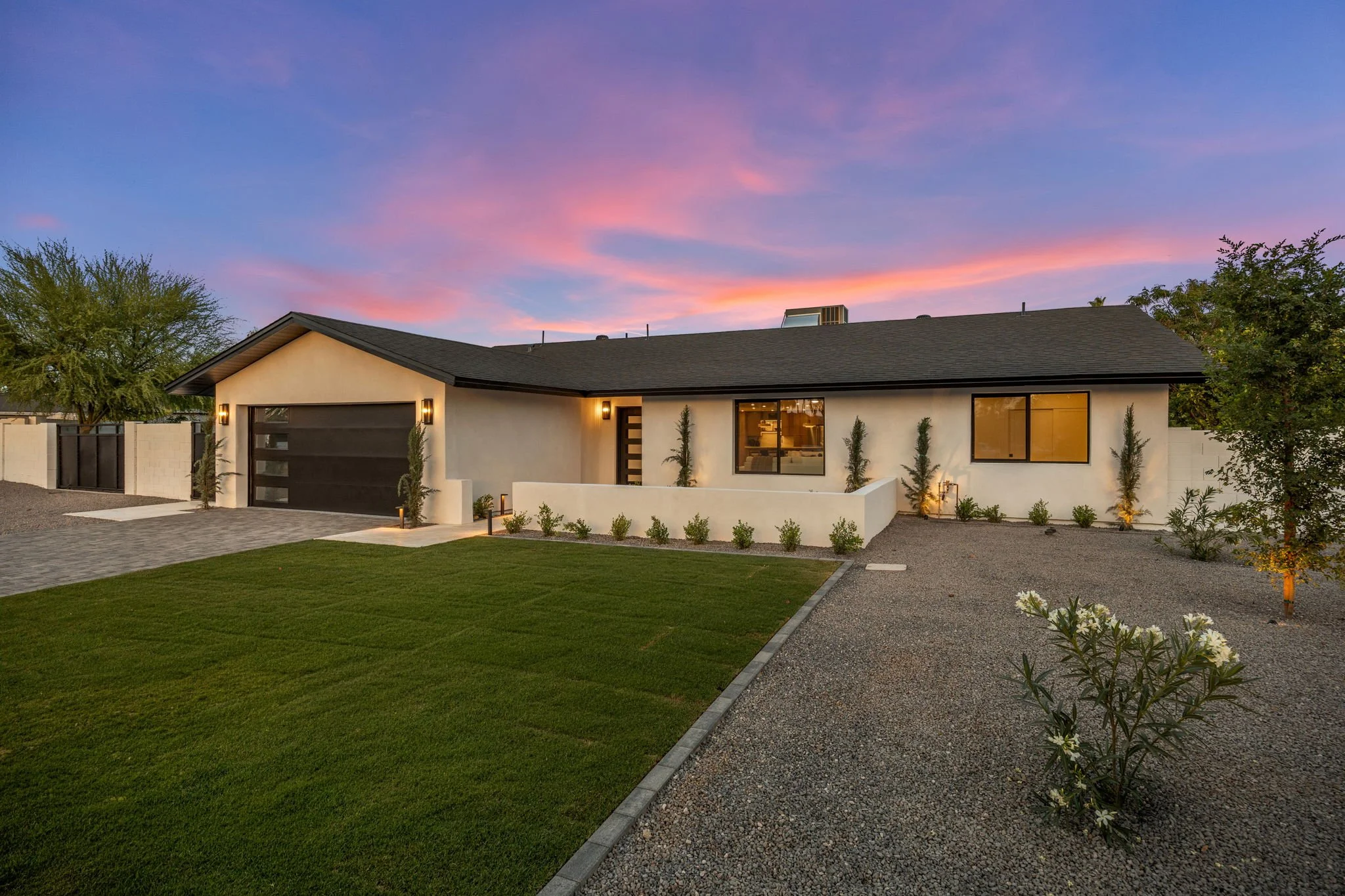 Modern single-story house with a dark garage door, large windows, and exterior lighting, surrounded by landscaped yard with grass, trees, and shrubs, under a colorful sky at sunset.