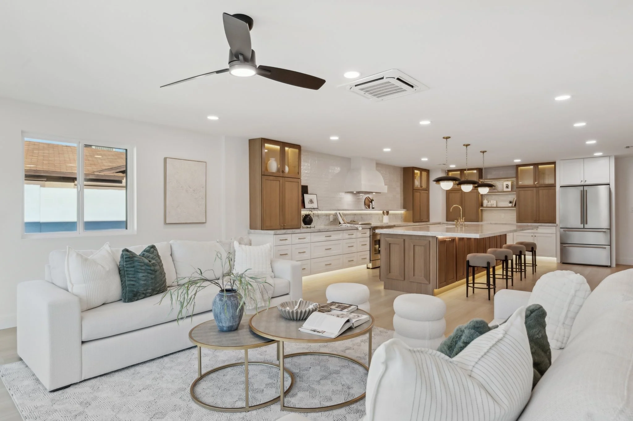 Modern open-concept living area and kitchen with white walls, large window, white seating, wooden cabinets, marble countertops, pendant and recessed lighting, ceiling fan, and bar stools.