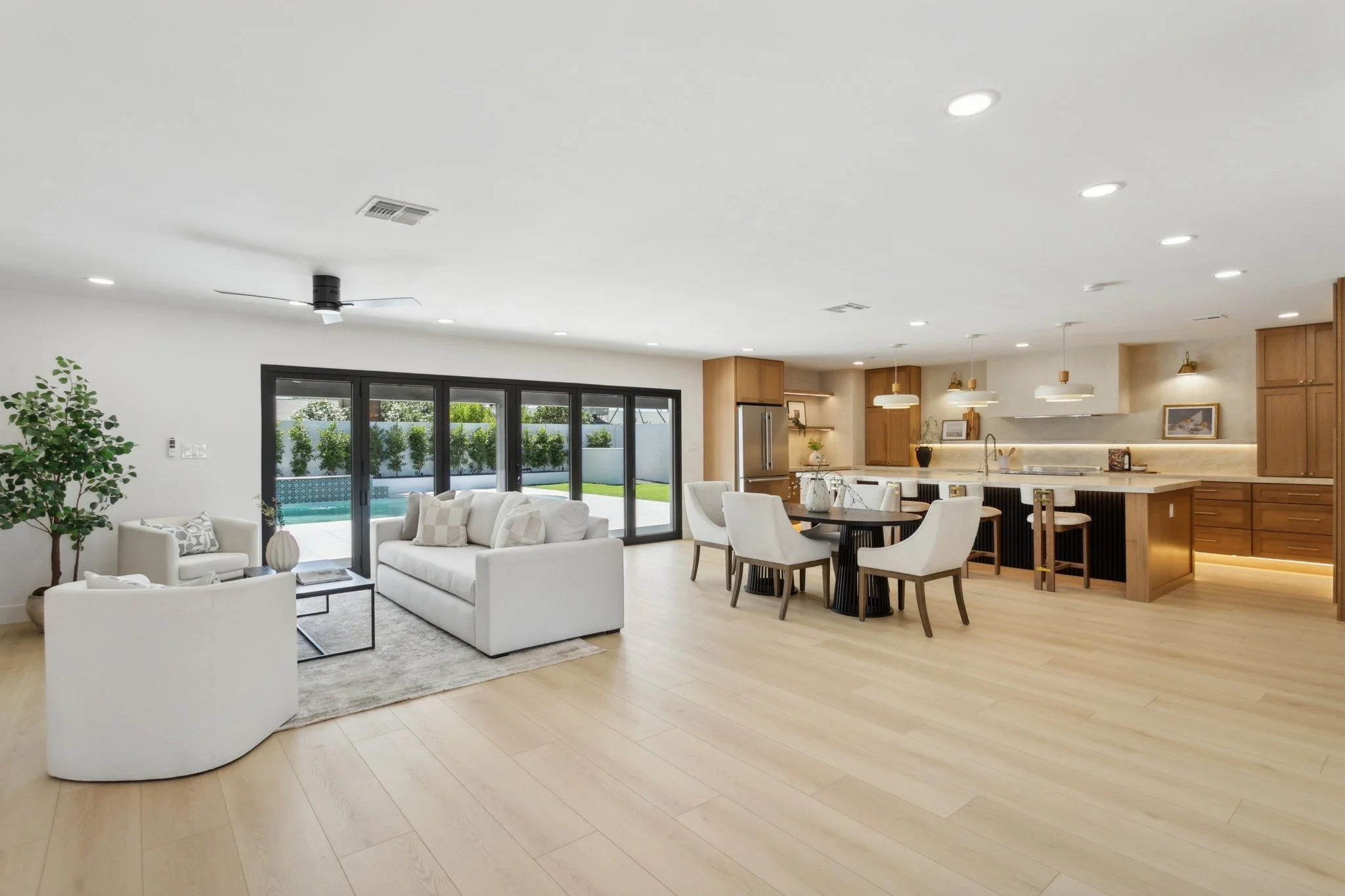 Modern open-concept living room and kitchen with white sofas, a dining table, kitchen island, wooden cabinetry, pendant lights, sliding glass doors, and a backyard with a lawn, fence, and pool.