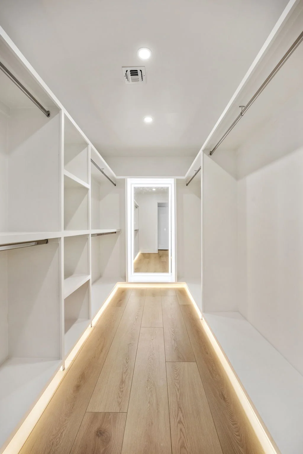 Empty walk-in closet with white shelves and hanging rods on both sides, wooden floor, and a full-length mirror at the end with LED lighting around it.